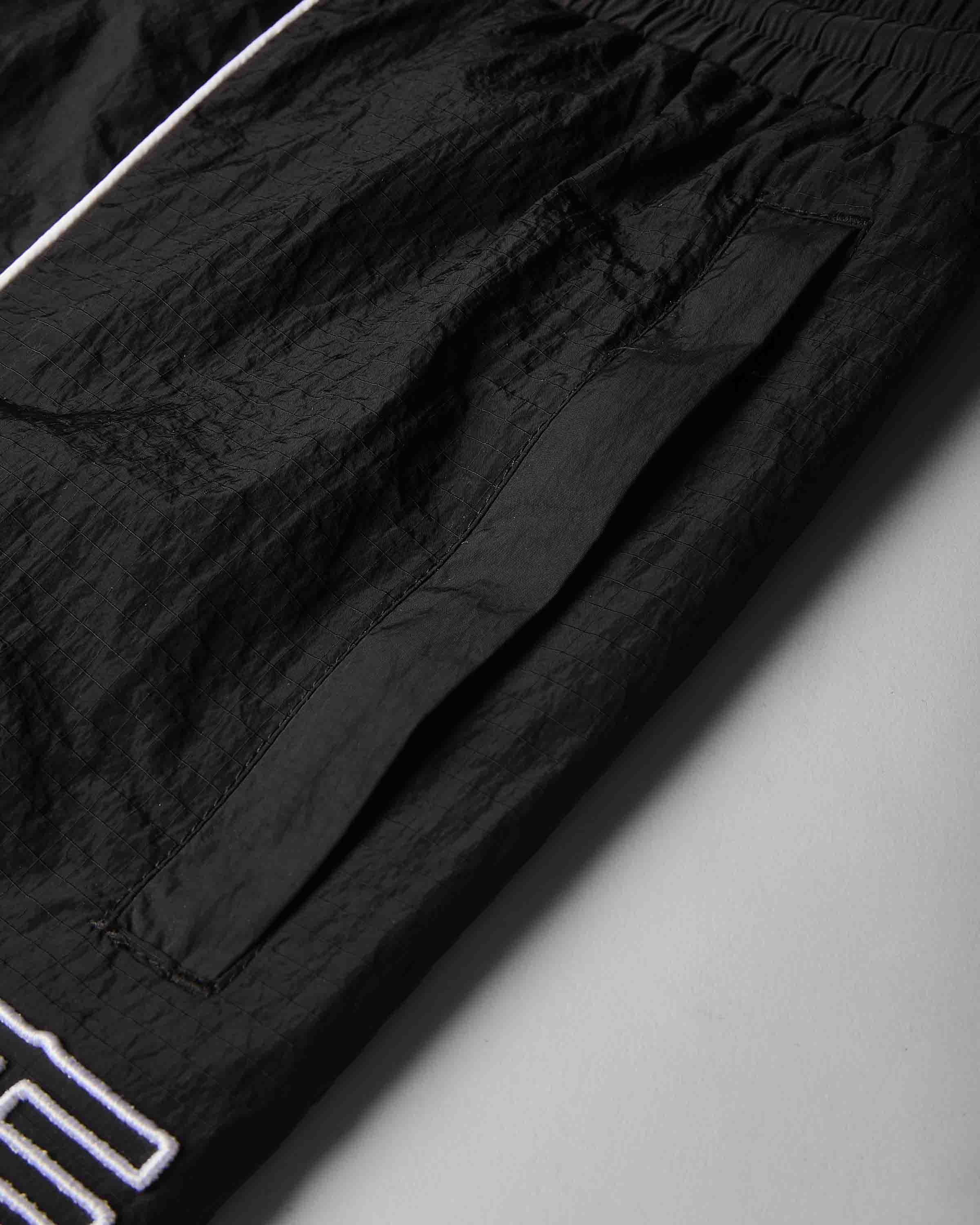 Close-up of black crinkled lightweight fabric with a white outlined logo at the bottom left, photographed on a light gray background.
