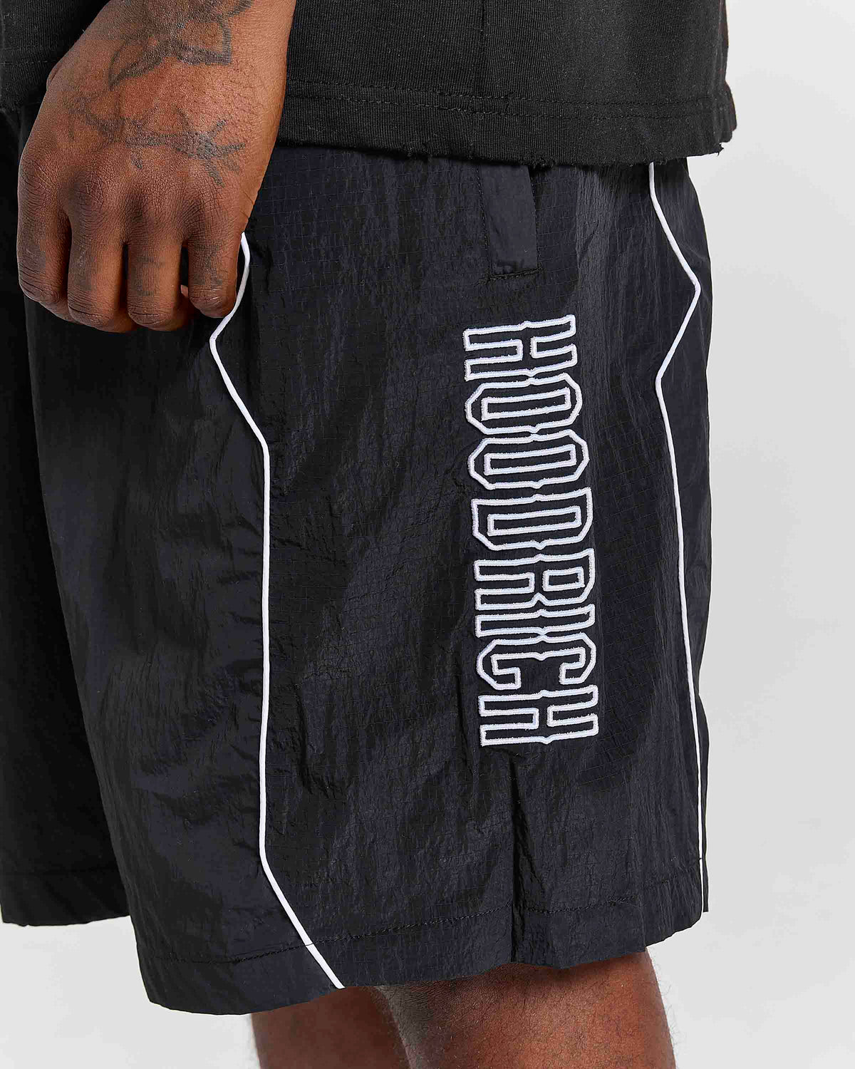 Close-up of a person wearing black athletic shorts with white piping and a large vertical white 'HOODRICH' logo on the side, paired with a black shirt against a light gray background.