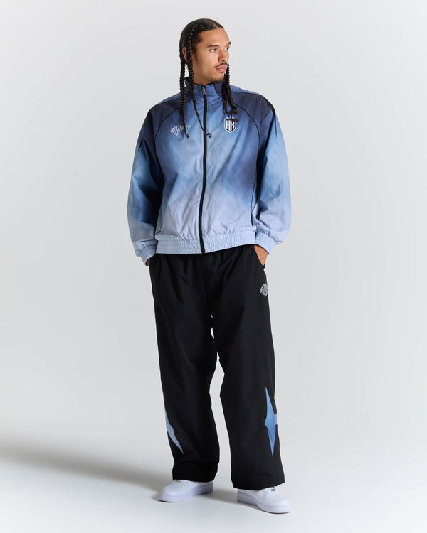A man modeling a light blue and navy gradient zip-up jacket with black track pants featuring light blue star details, set against a plain light gray studio background.