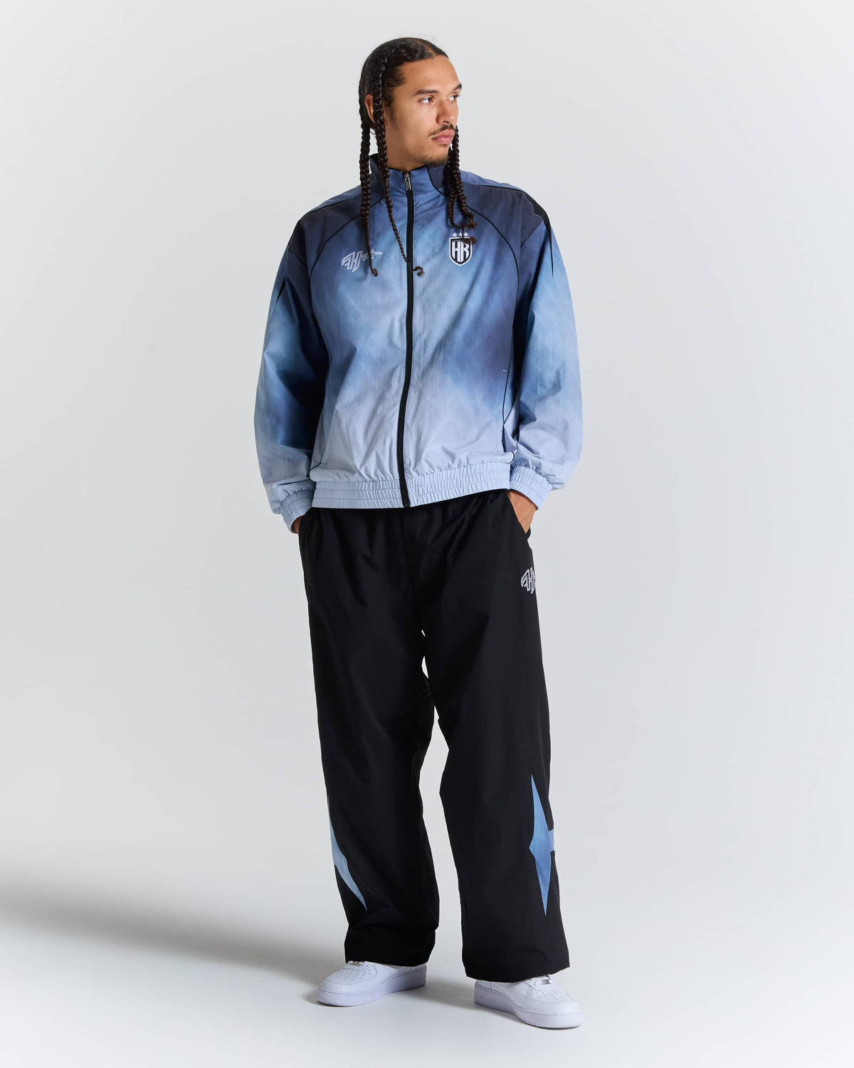 A man modeling a light blue and navy gradient zip-up jacket with black track pants featuring light blue star details, set against a plain light gray studio background.