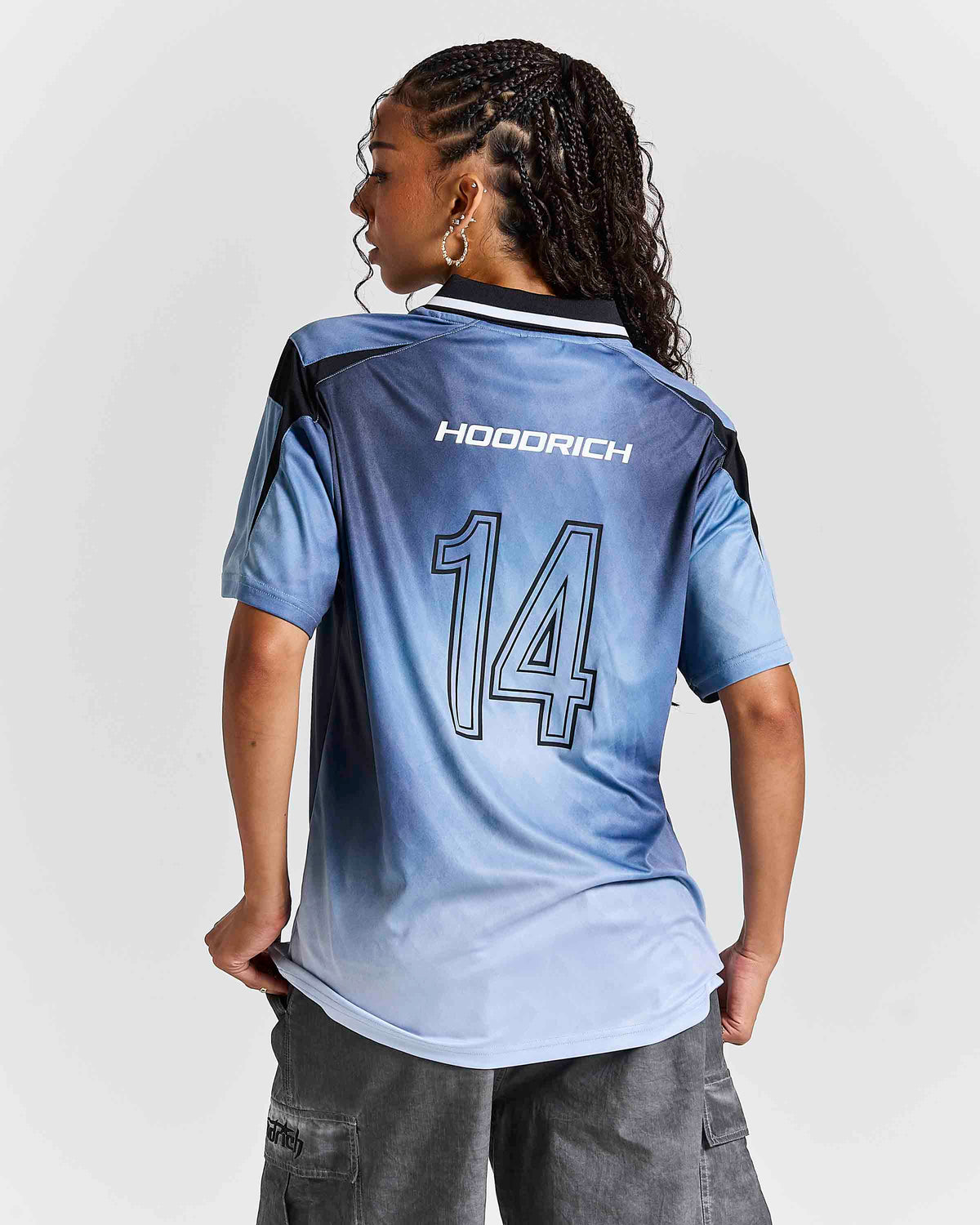 A woman shown from the back wearing a light blue and white gradient HOODRICH soccer jersey with the number 14 on the back, paired with gray cargo pants against a light gray studio background.
