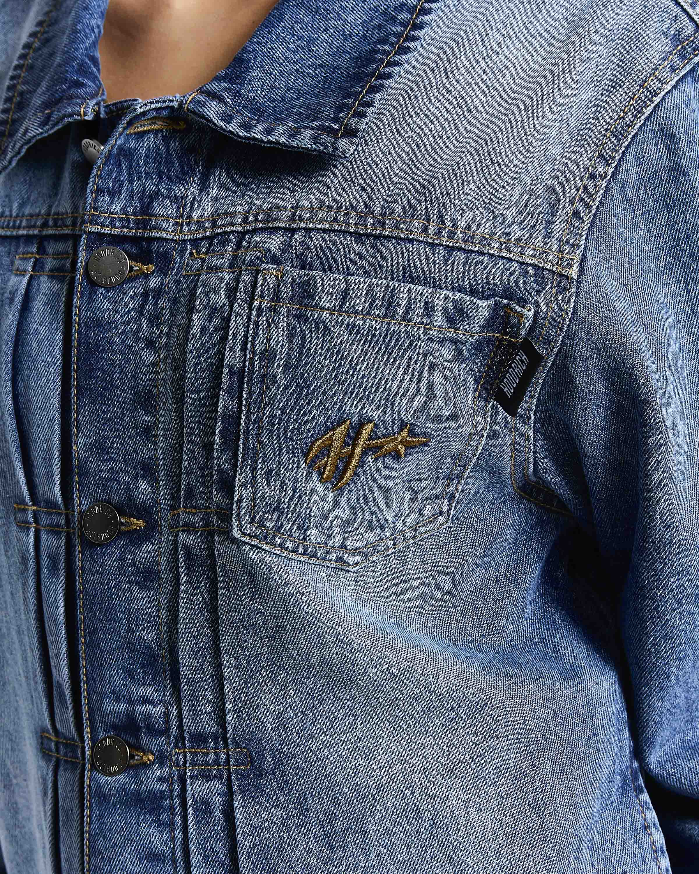 Close-up of a blue denim jacket featuring a chest pocket with a gold embroidered logo and dark metal snap buttons.