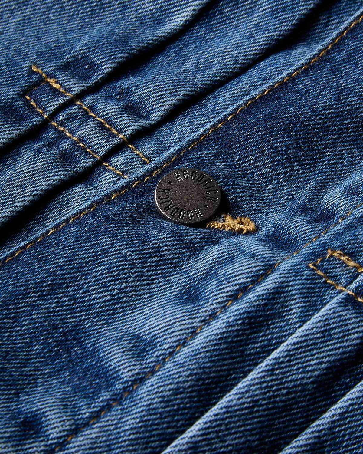 Close-up of a dark metal button on a blue denim garment, showing the textured fabric weave and contrast stitching.