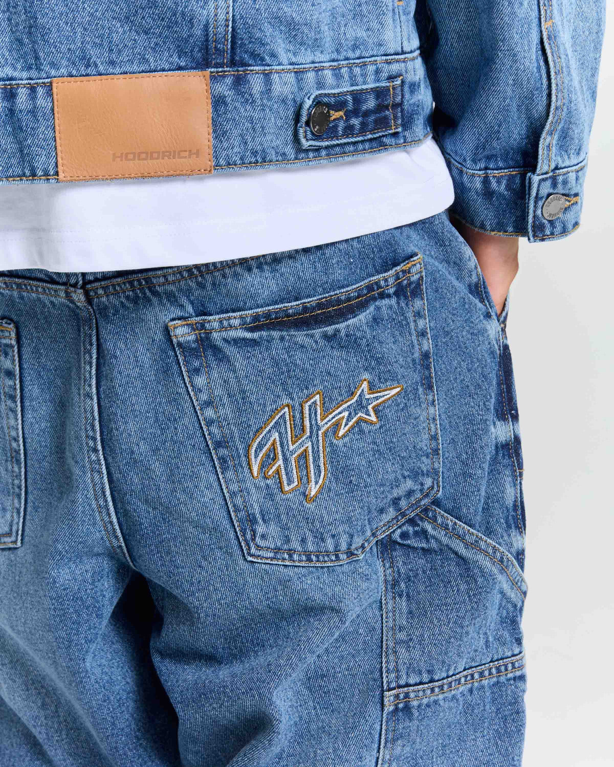 Close-up of a person wearing blue denim jeans with a white t-shirt underneath and a matching blue denim jacket. The back pocket of the jeans features a gold embroidered monogram logo, and a tan leather brand patch is visible on the jacket.