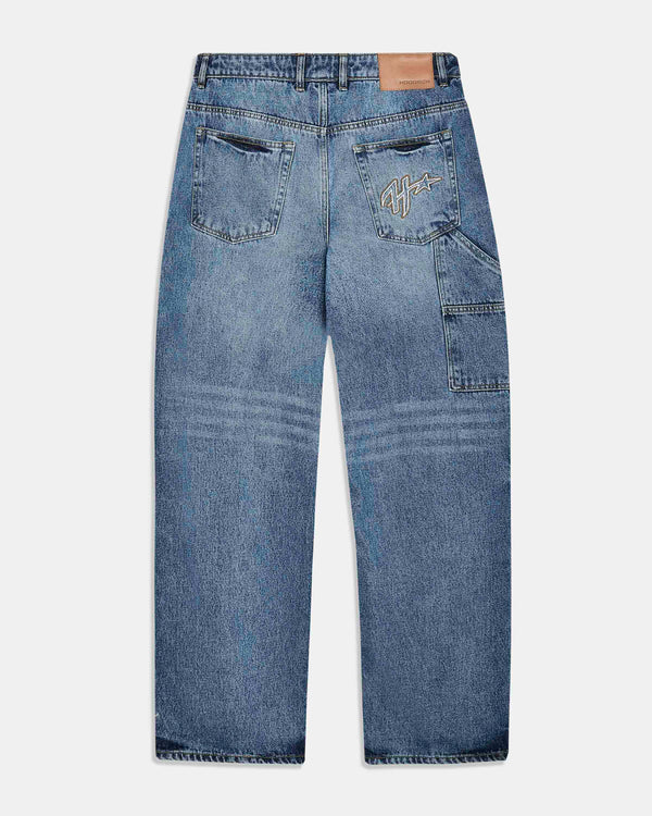 Back view of a pair of blue denim jeans with a light wash, featuring a tan leather patch on the back waistband and a stylized embroidered logo on the right back pocket.
