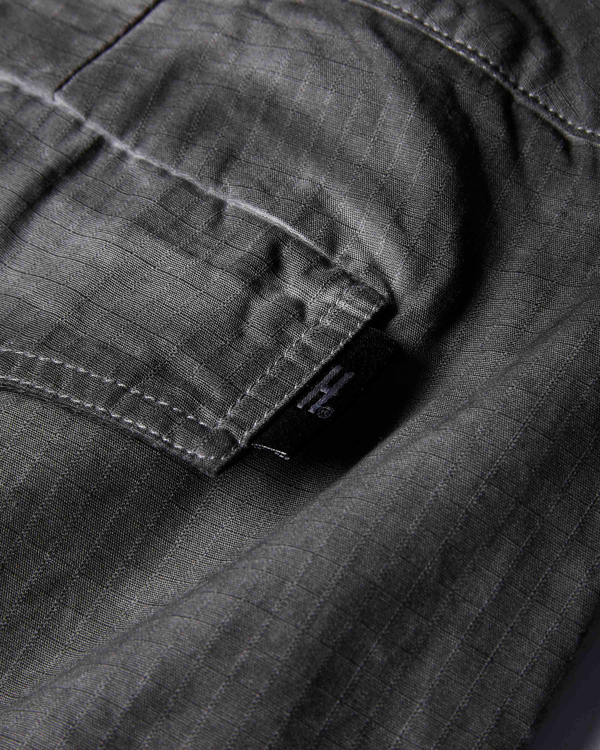 Close-up of dark gray textured fabric with a pocket and a small black brand tab showing the stylized H logo.