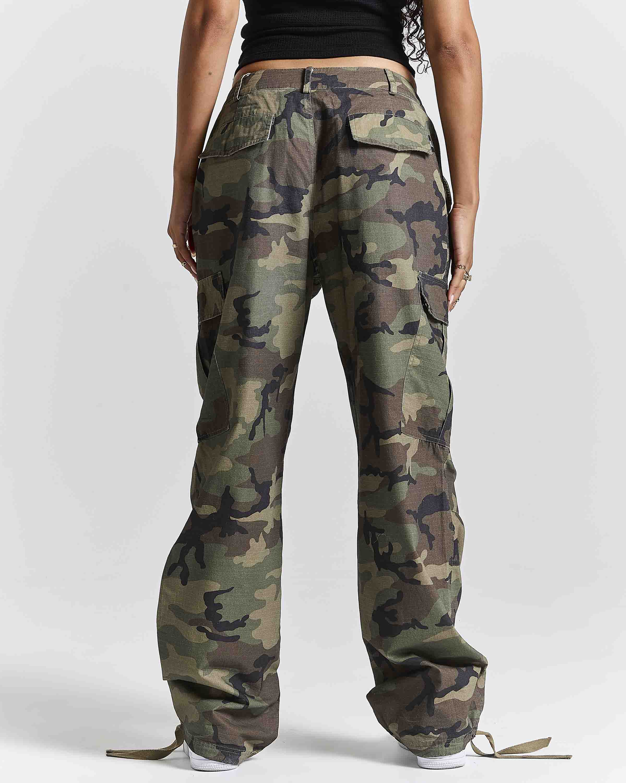 A person shown from the back wearing loose camouflage cargo pants with a black top and white sneakers, photographed against a light gray studio background.