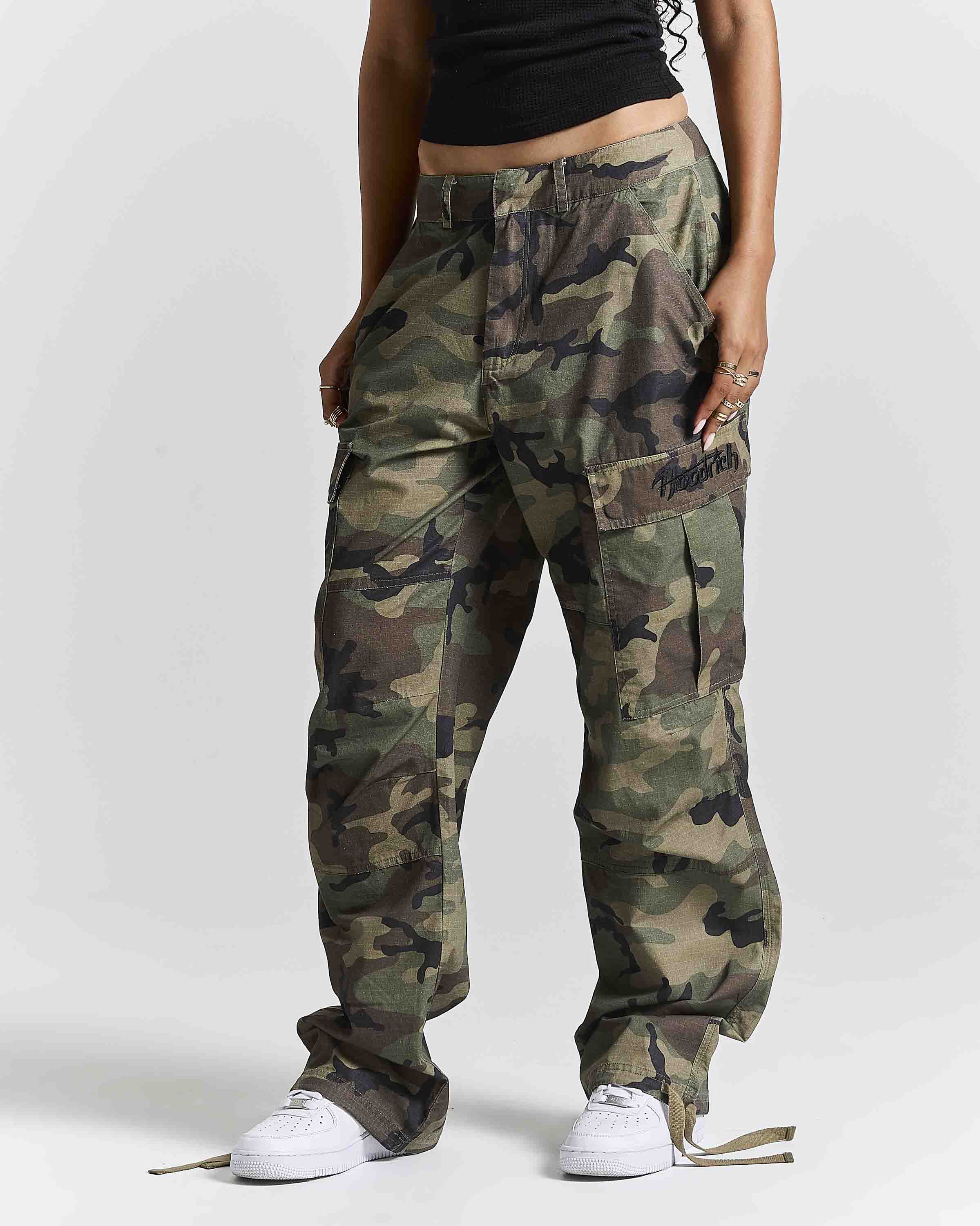 A person wearing loose-fit camouflage cargo pants with a black cropped top and white sneakers, standing against a light gray studio background.