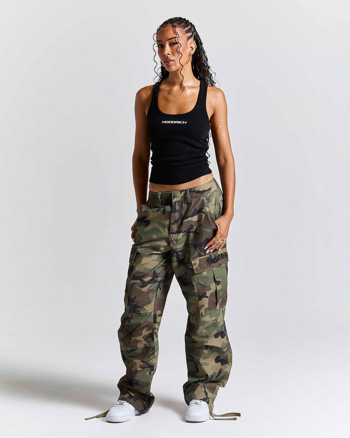 A model stands against a light gray studio background wearing a black tank top with white text, loose camouflage cargo pants, and white sneakers. The tank top has the brand name 'HOODRICH' printed on the chest.