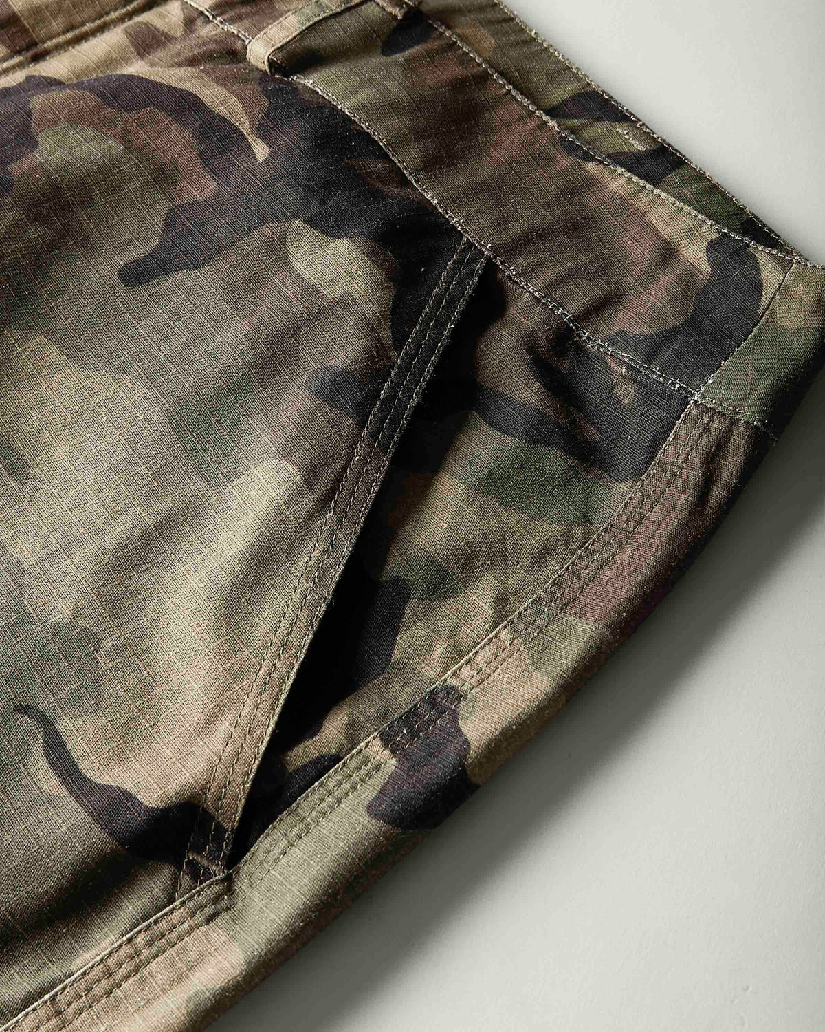 Close-up of camouflage pants showing the fabric pattern and side seam details against a light gray background.