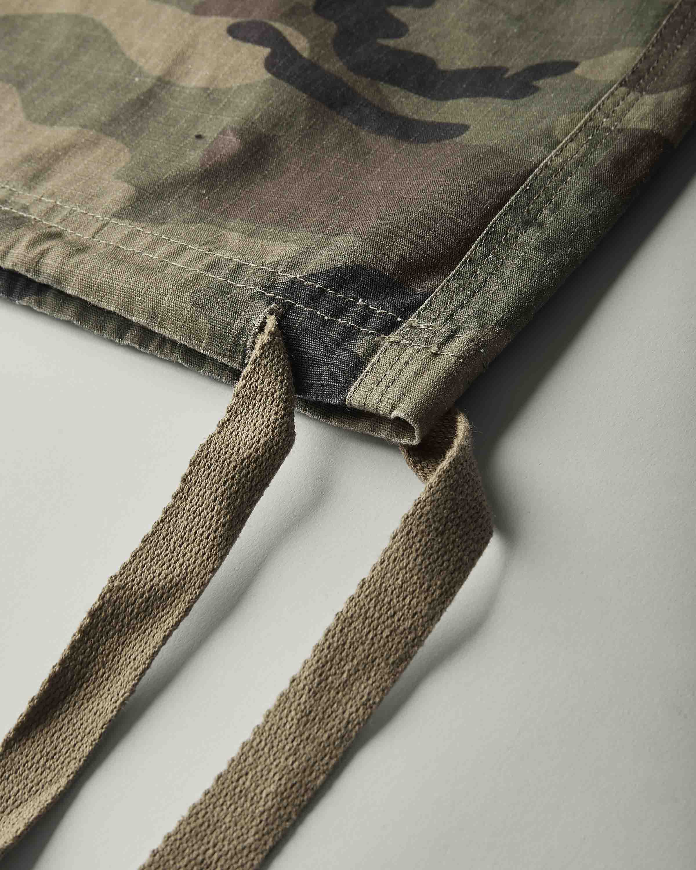 Close-up of a camouflage fabric bag corner with tan woven drawstring straps, photographed on a light gray background.