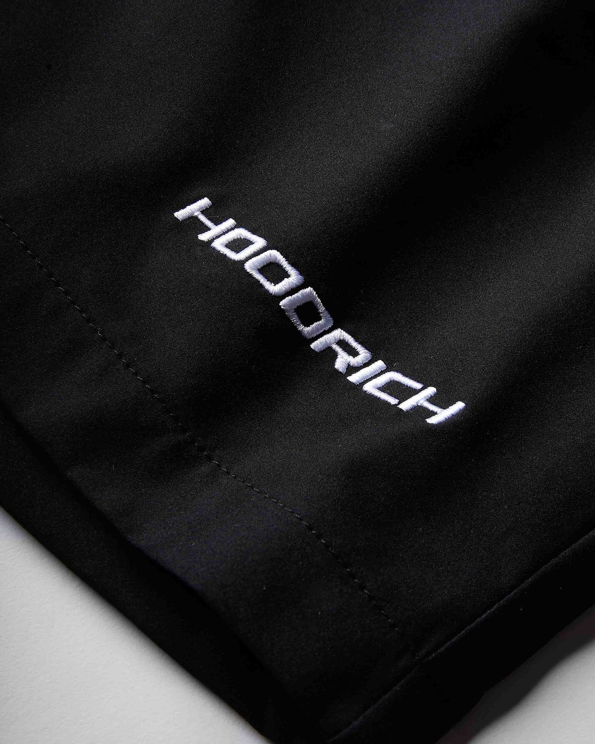 Close-up of a black athletic garment, likely shorts, featuring the brand name 'HOODRICH' embroidered in white with a silver outline on the side.