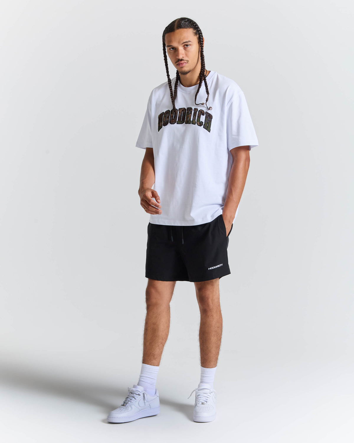 A man modeling a white short-sleeve t-shirt with 'HOODRICH' printed in black across the chest, paired with black athletic shorts, white socks, and white sneakers, against a light gray studio background.
