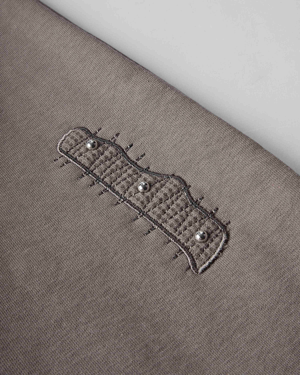 Close-up of a taupe knit garment featuring a decorative embroidered patch with three silver-tone studs.