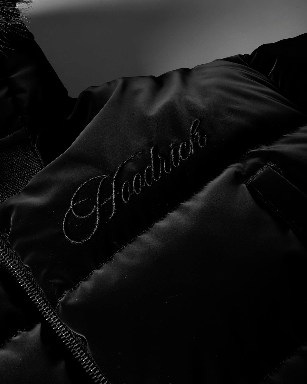 Close-up of a black puffer jacket with a subtle embroidered script logo reading 'Hodrich' on the chest.
