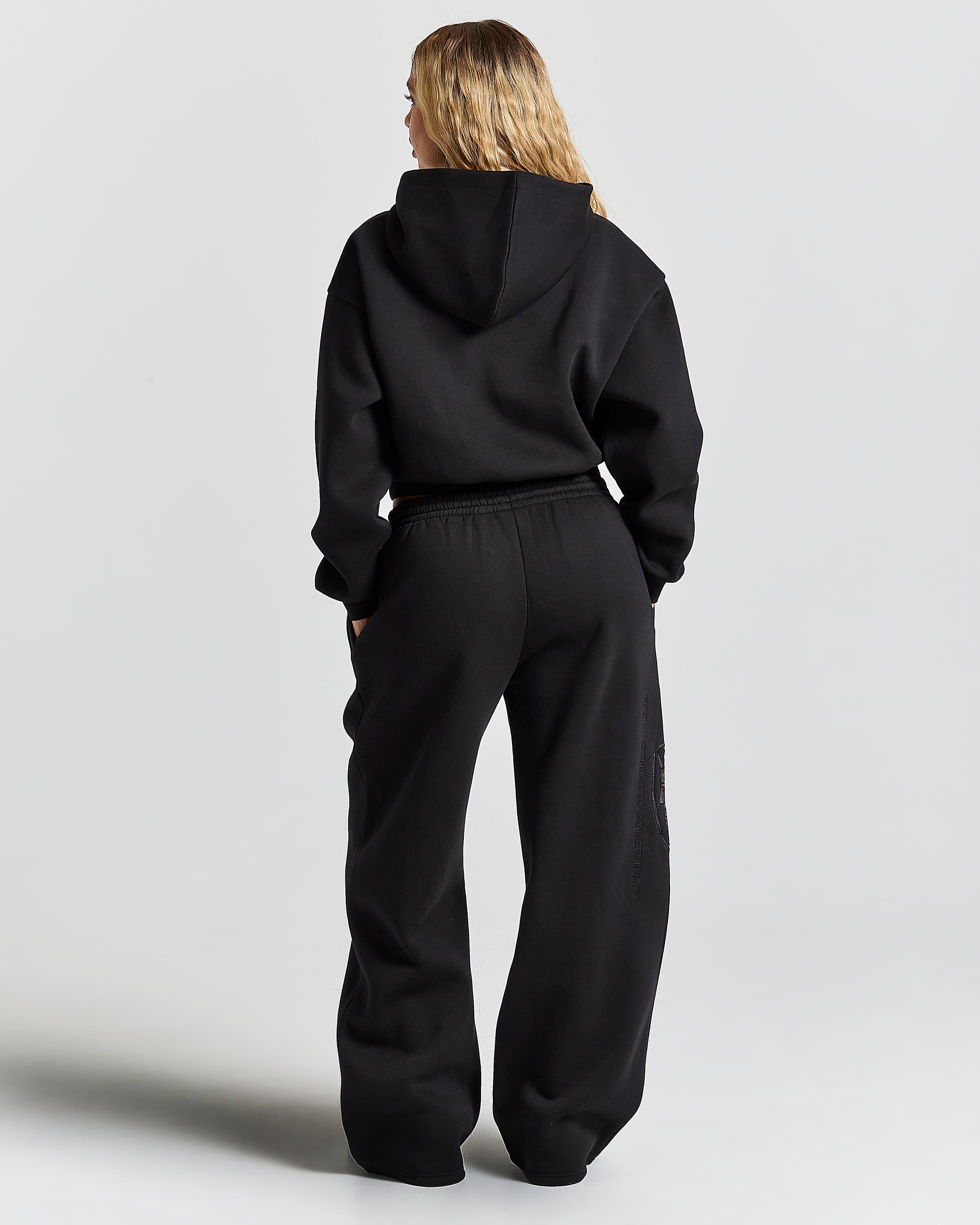 A woman viewed from behind wearing a black cropped hoodie and matching black wide-leg sweatpants against a light gray studio background.