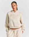 A model wearing a cream-colored cropped hoodie with 'HOODRICH' embossed on the chest, paired with matching jogger pants, against a light gray studio background.