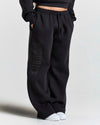 Black wide-leg sweatpants with a tonal vertical 'HOODRICH' logo on the left leg, shown on a model wearing white sneakers against a light gray background.