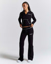A woman modeling a black athletic tracksuit with white contrast stitching and white logo text on the chest and thigh, photographed against a light gray studio background.