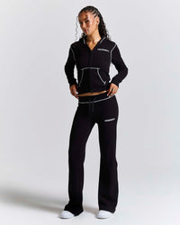 A woman modeling a black athletic tracksuit with white contrast stitching and white logo text on the chest and thigh, photographed against a light gray studio background.