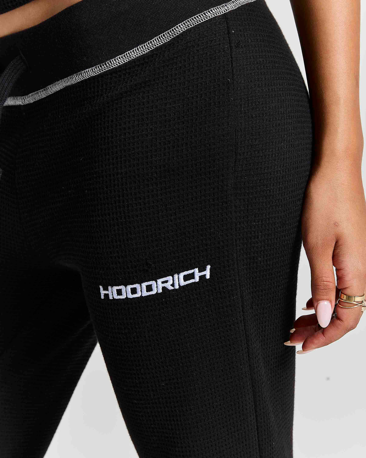 Close-up of a person wearing black textured hooded sweatpants with a white HOODRICH logo on the thigh and a shimmering waistband.