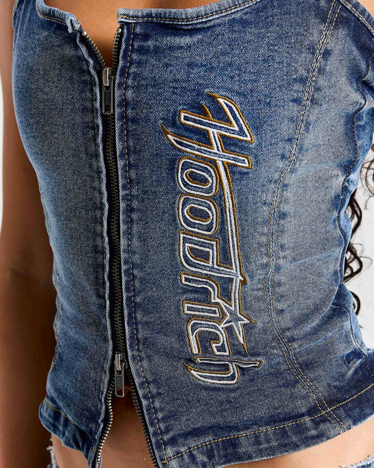 Close-up of a blue denim zip-front top with large metallic embroidered 'Hooded' lettering on the front, worn by a model.