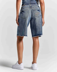 A person shown from the back wearing medium-wash blue denim shorts with a raw frayed hem, paired with white sneakers against a light gray studio background.