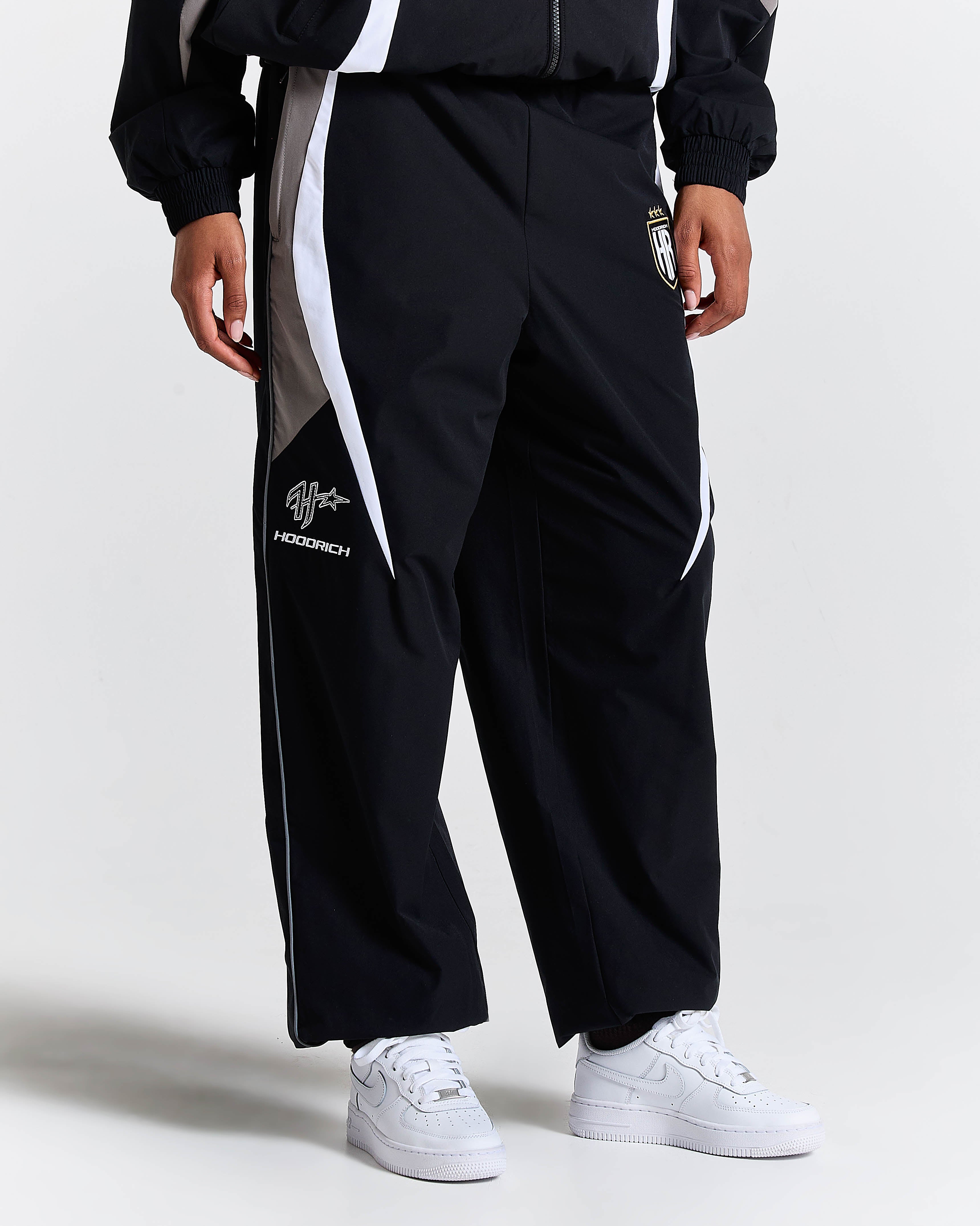 Person wearing black tracksuit pants with white stripes and white sneakers on a light gray background