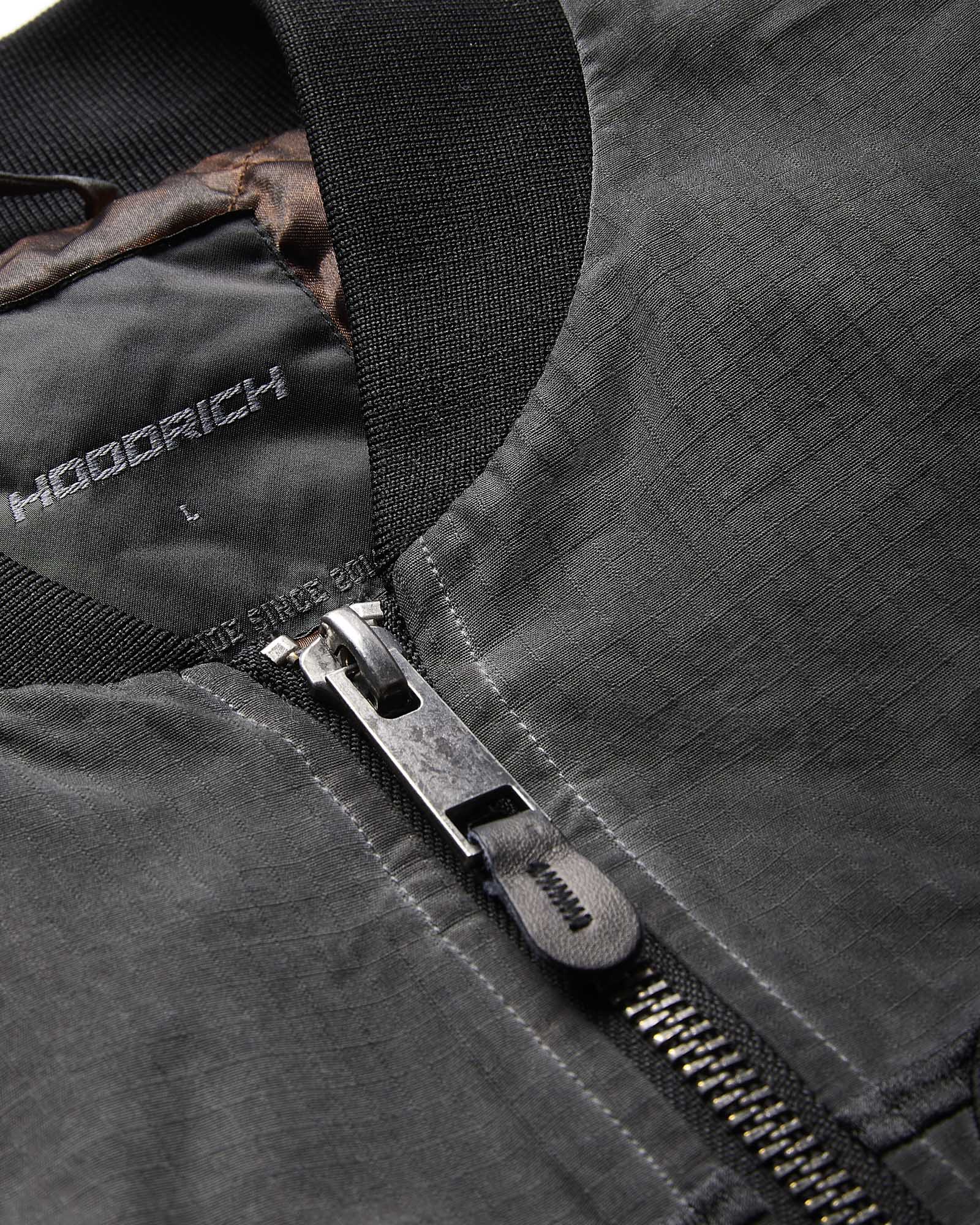 Close-up of a dark gray zippered jacket with a black ribbed collar and a silver metal zipper pull. The brand name 'HOODRICH' is printed on the inner neck lining.