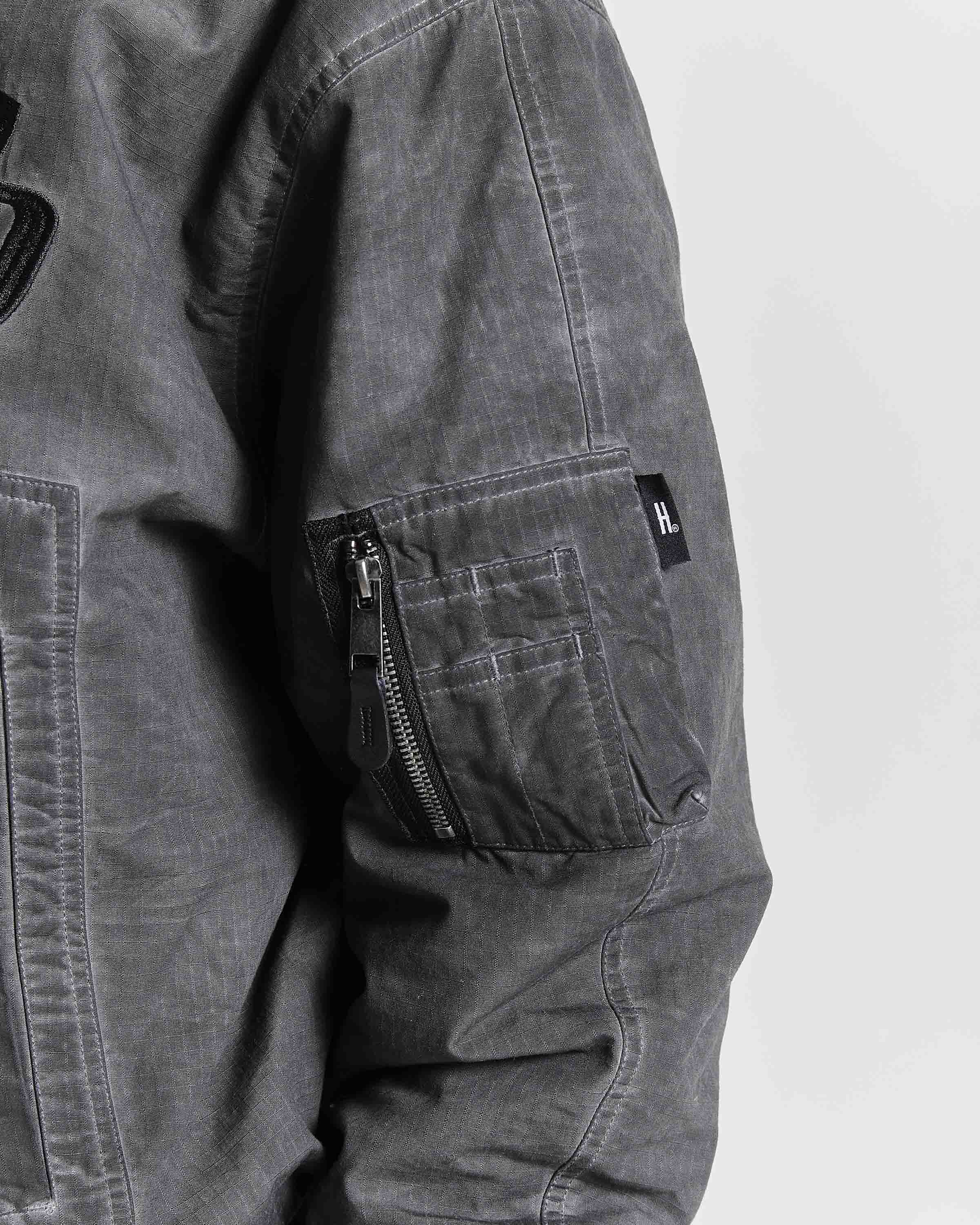 Close-up of a dark gray washed canvas utility jacket, showing a zippered cargo pocket on the upper arm with a small black brand tab.