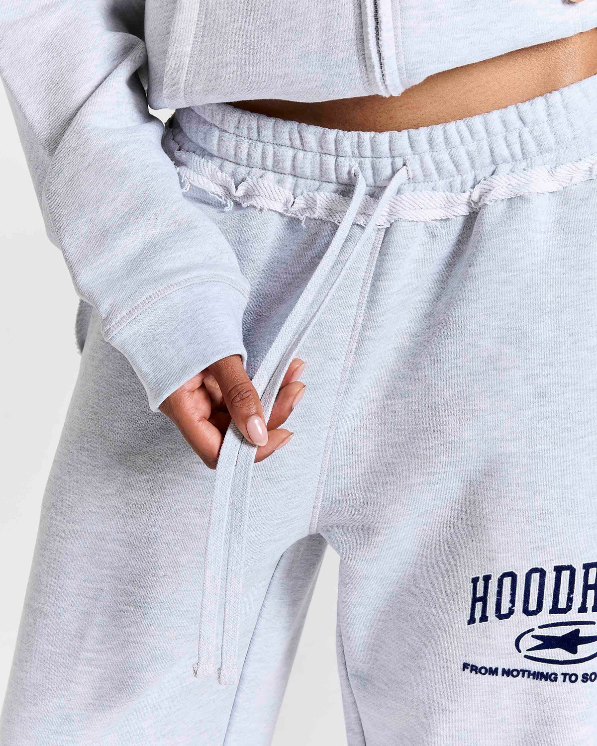 Close-up of a person wearing light heather gray sweatpants with a drawstring waist and a navy graphic print on the left leg that reads 'HOODRICH'