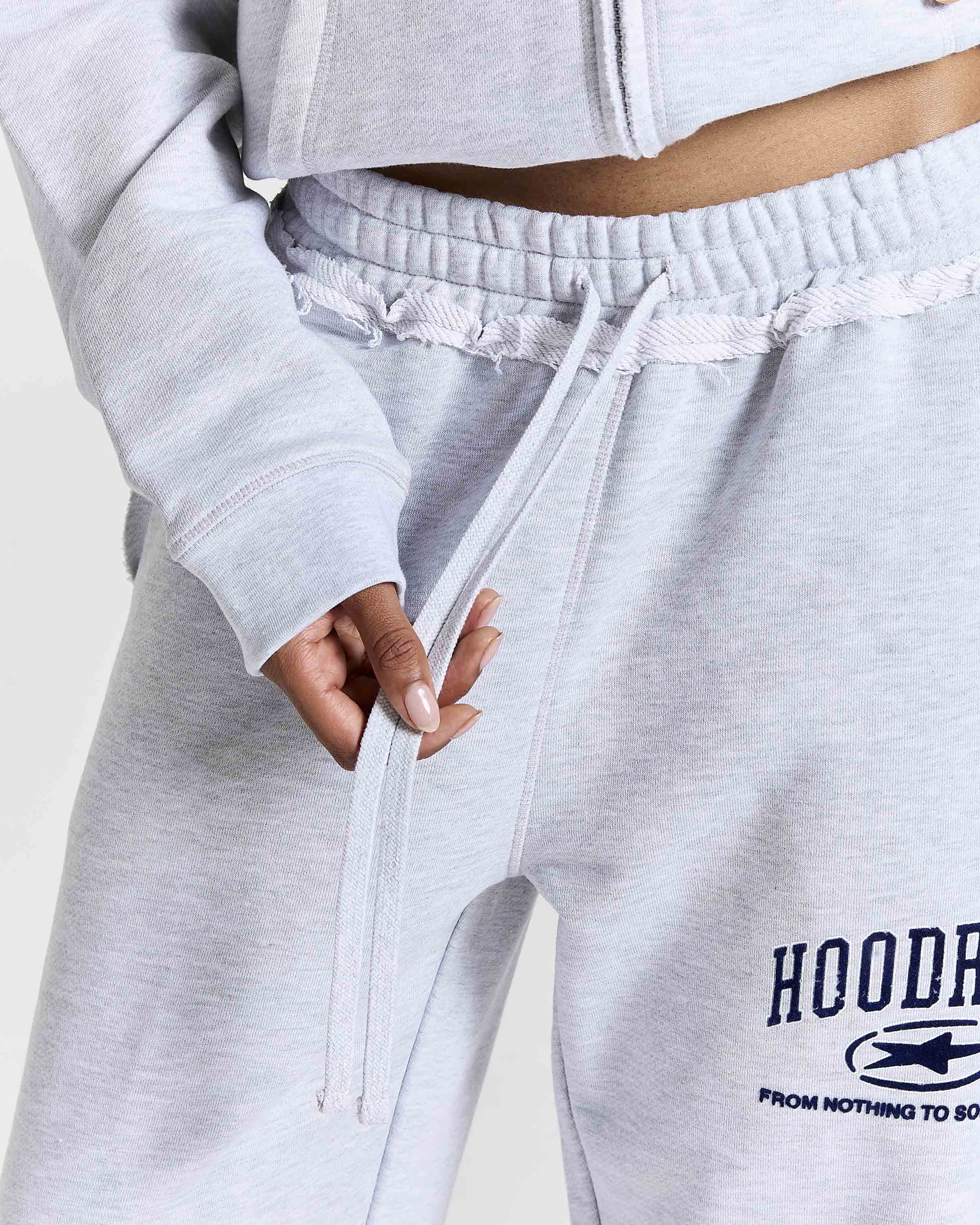 Close-up of a person wearing light heather gray sweatpants with a drawstring waist and a Navy graphic print on the left leg that reads 'HOODRICH' with a small logo and 'FROM NOTHING TO SOMETHING'.