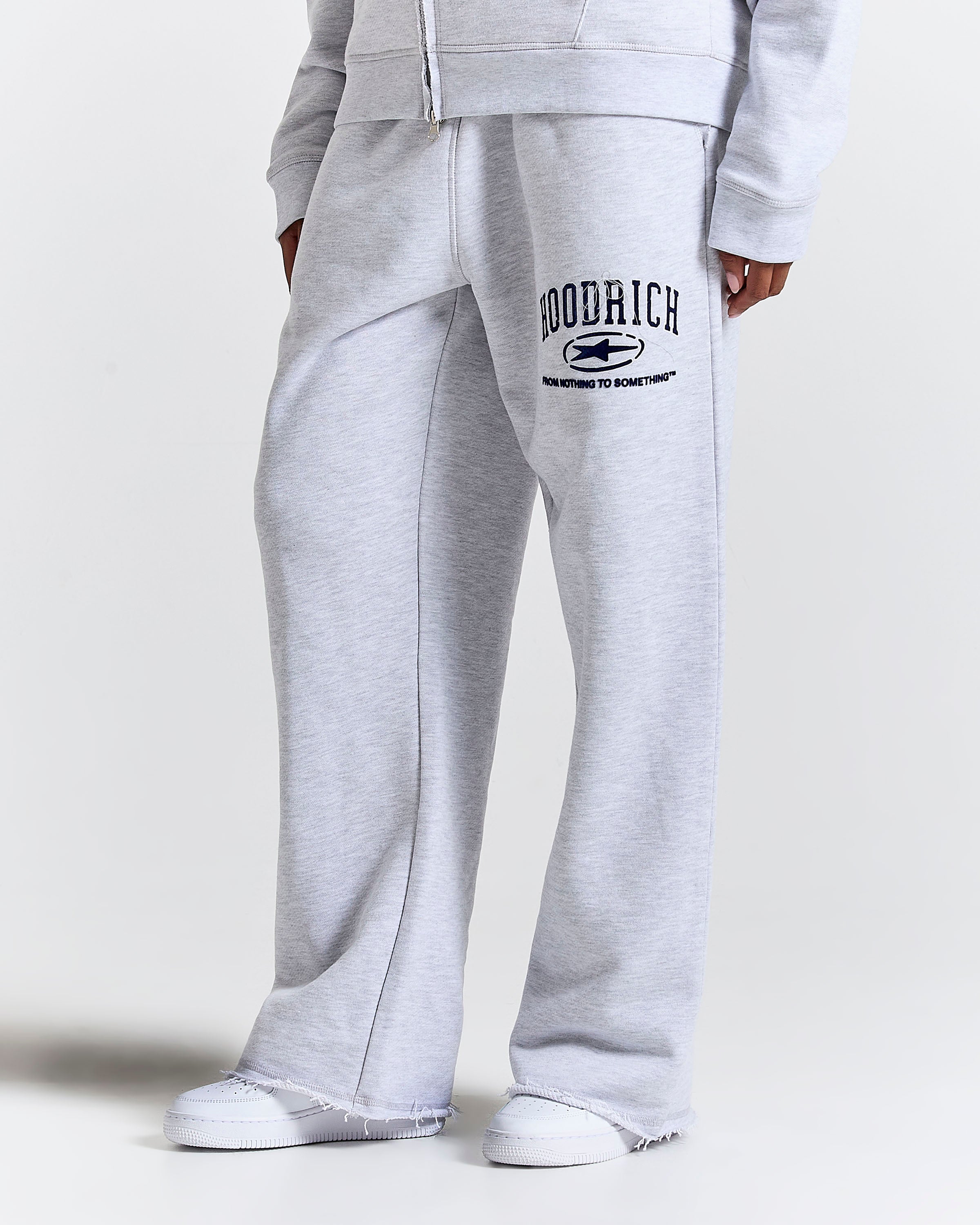 Close-up of a person wearing light heather gray sweatpants with a drawstring waist and a Navy graphic print on the left leg that reads 'HOODRICH' with a small logo and 'FROM NOTHING TO SOMETHING'.
