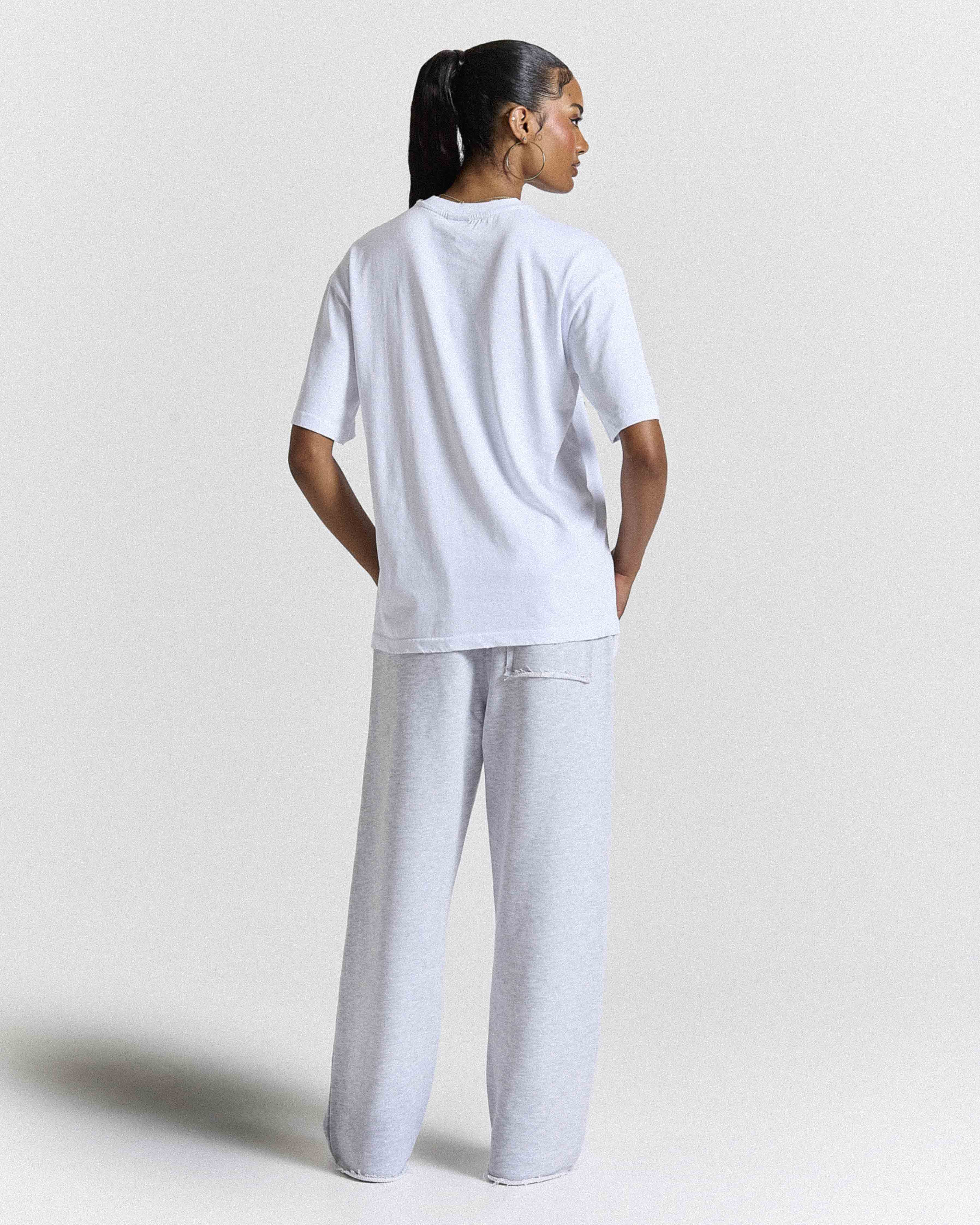 Back view of a female model wearing a white t-shirt and grey joggers on a grey studio backdrop.