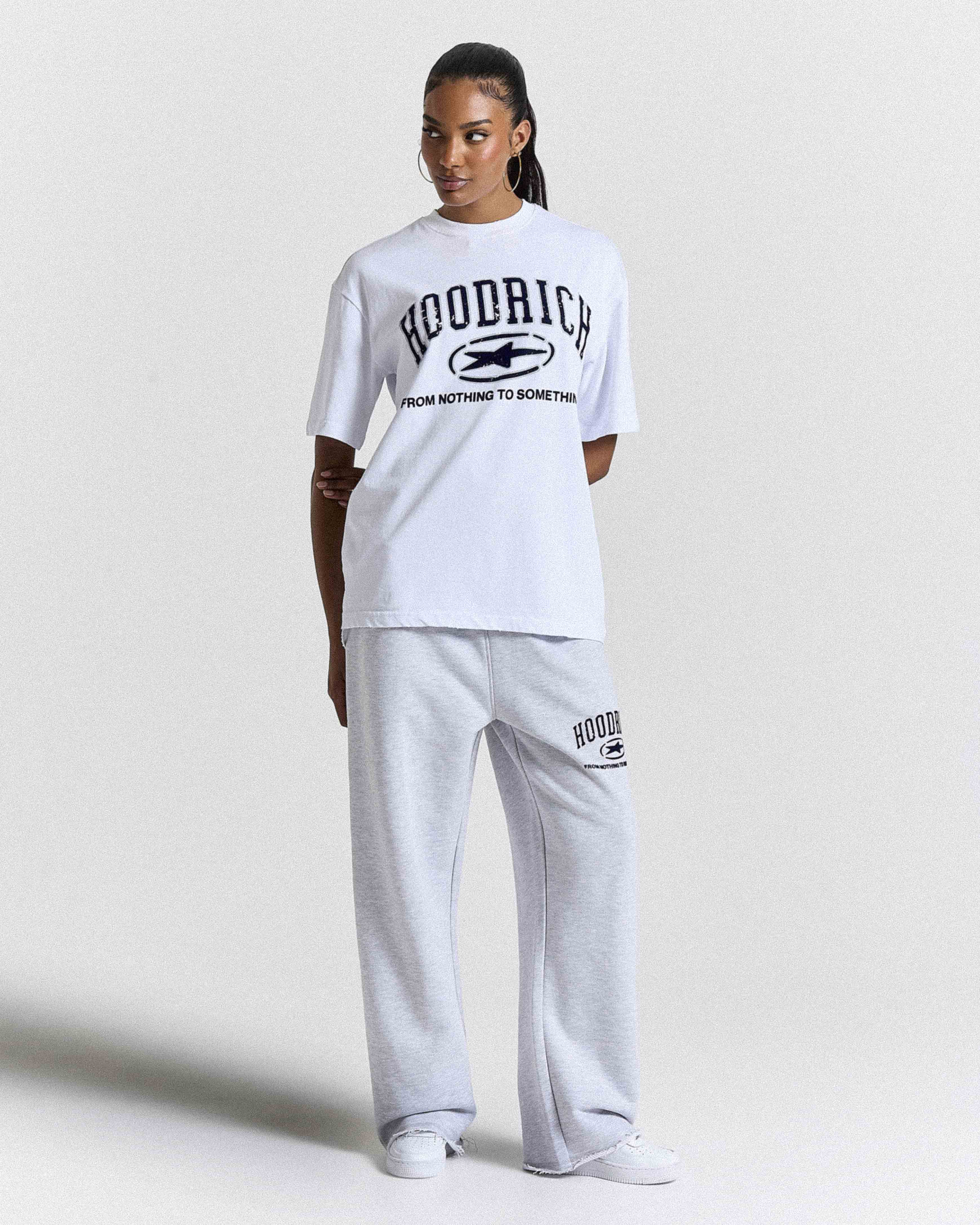 A model wearing a white short-sleeve graphic t-shirt and light gray sweatpants with black 'HOODRICH' branding, standing against a light gray studio background.