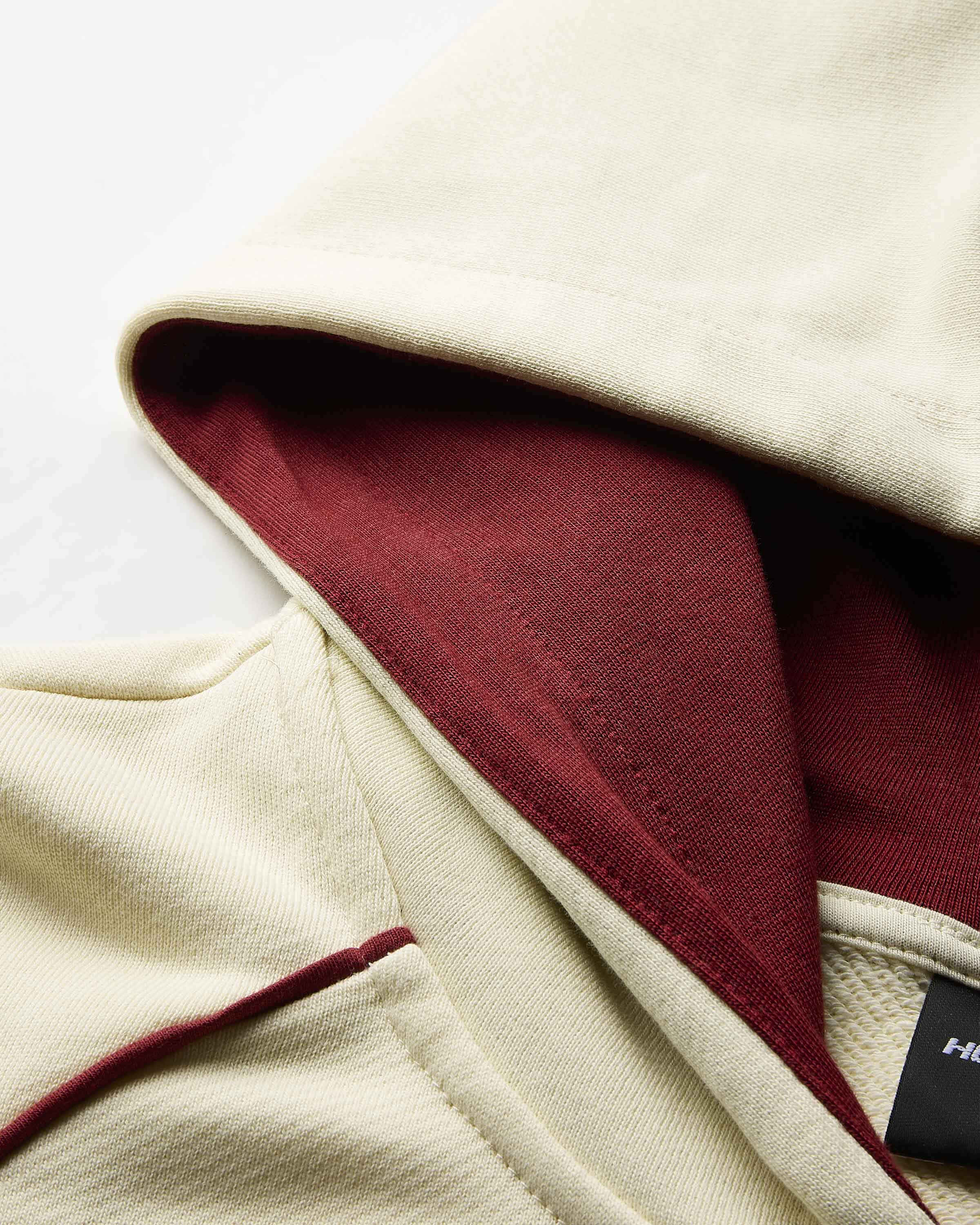 Close-up detail of a cream-colored hooded garment with a contrasting dark red inner lining and a small black brand tag.