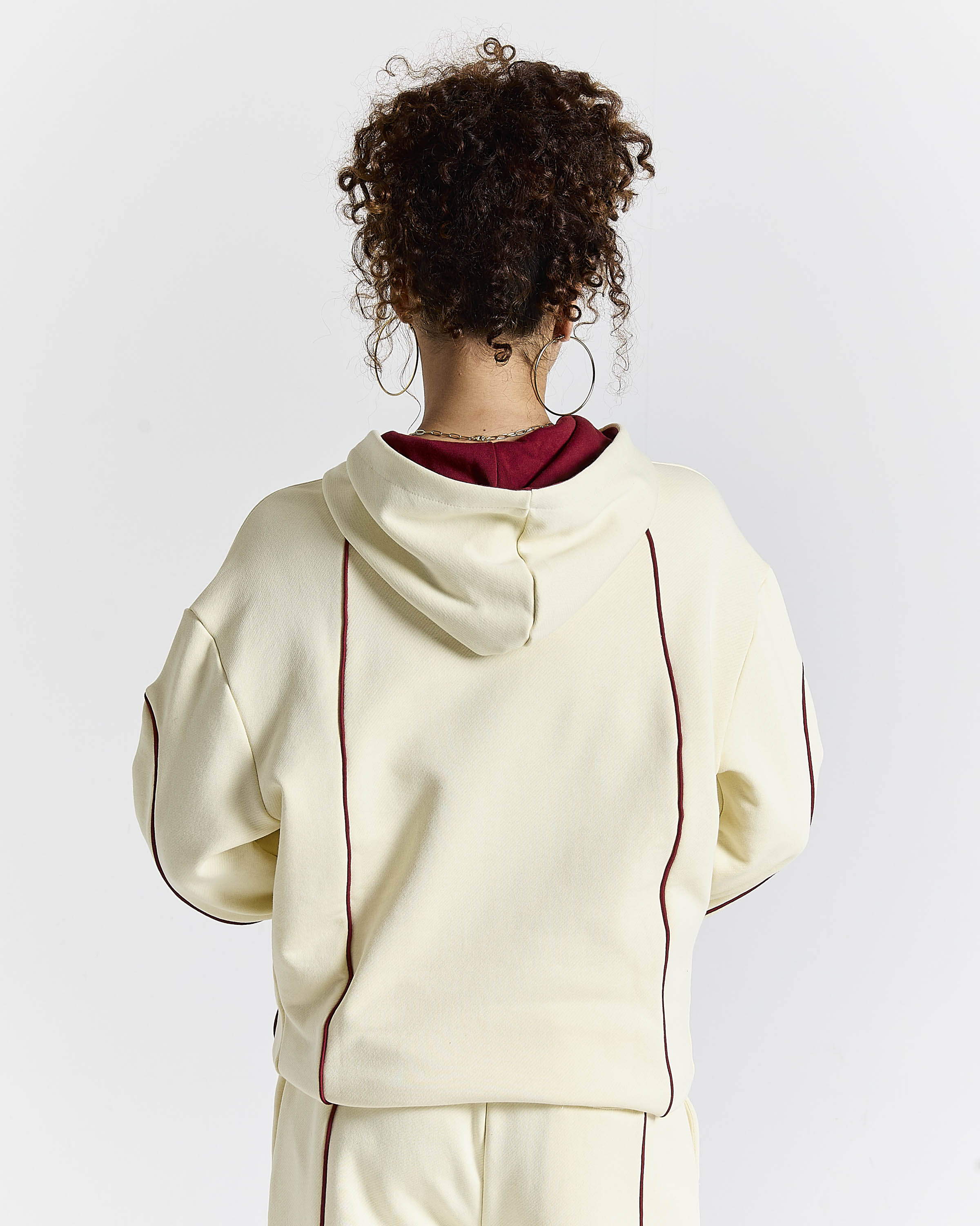 A person shown from the back wearing a cream-colored hoodie with dark red contrast piping and a dark red inner hood lining, paired with matching cream pants against a light gray studio background.