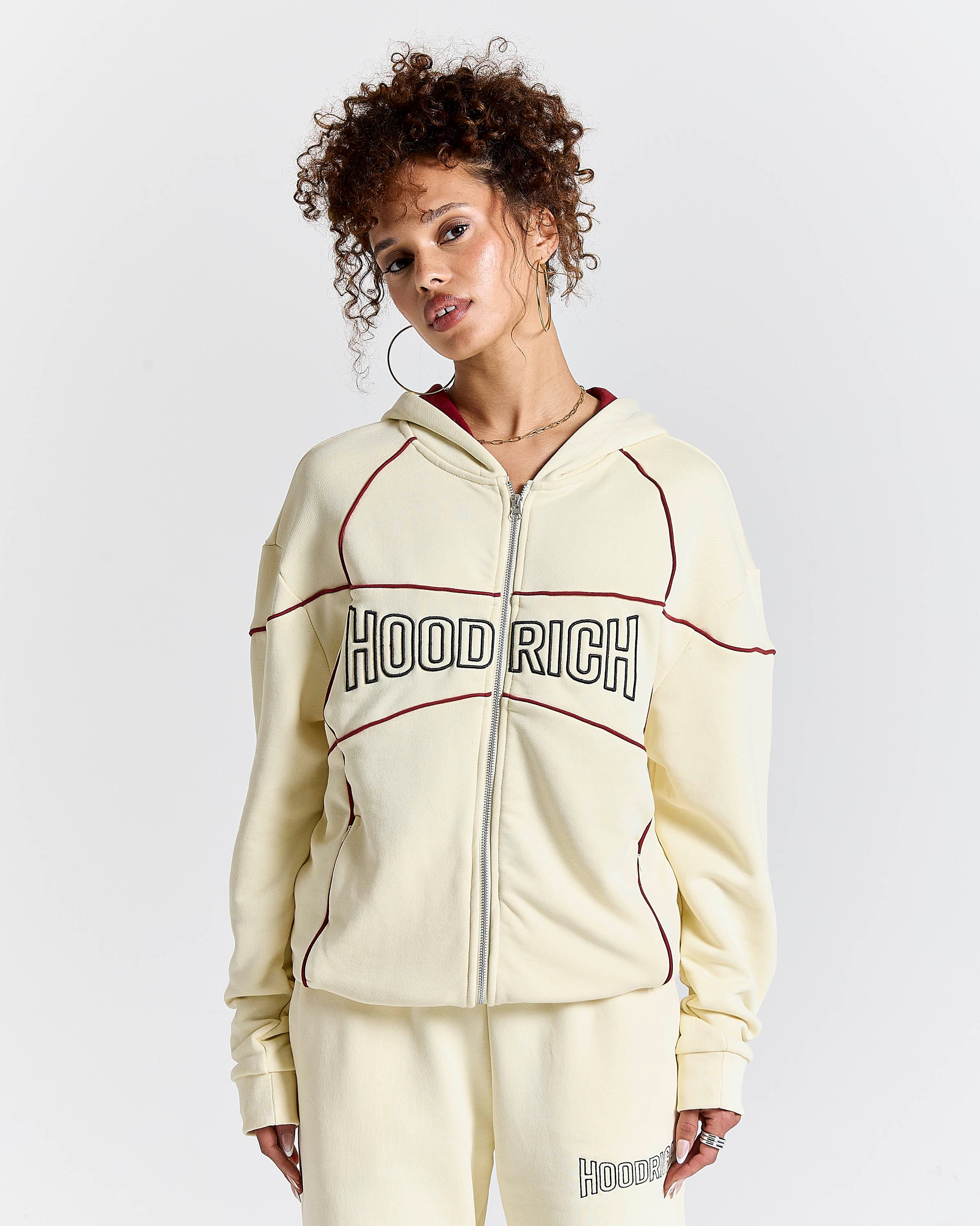A model wearing a cream-colored zip-up hoodie with 'HOODRICH' printed across the chest in black and white, accented with thin red lines, paired with matching sweatpants against a light gray background.
