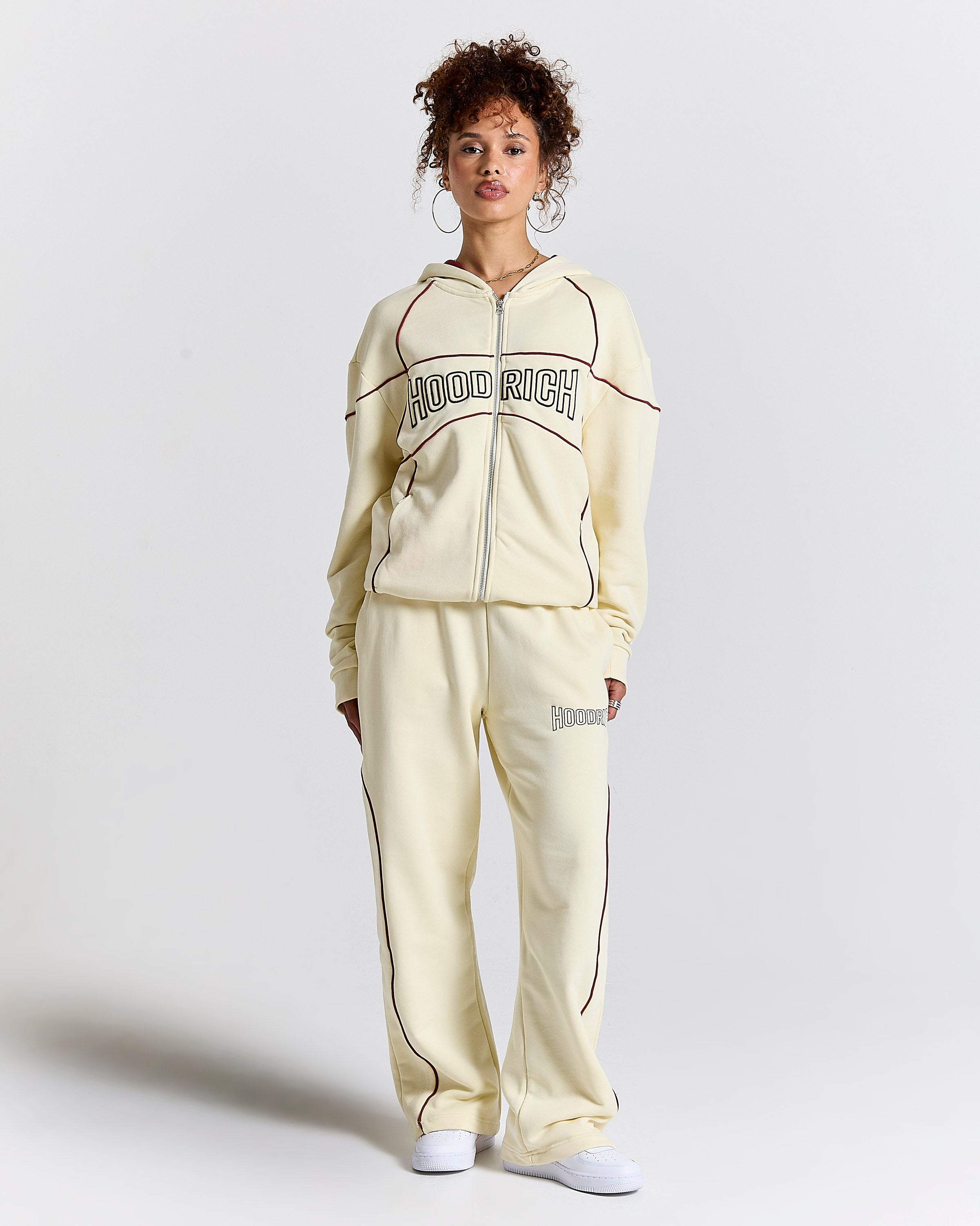 Woman wearing a cream-colored 'Hood Rich' tracksuit on a light gray background