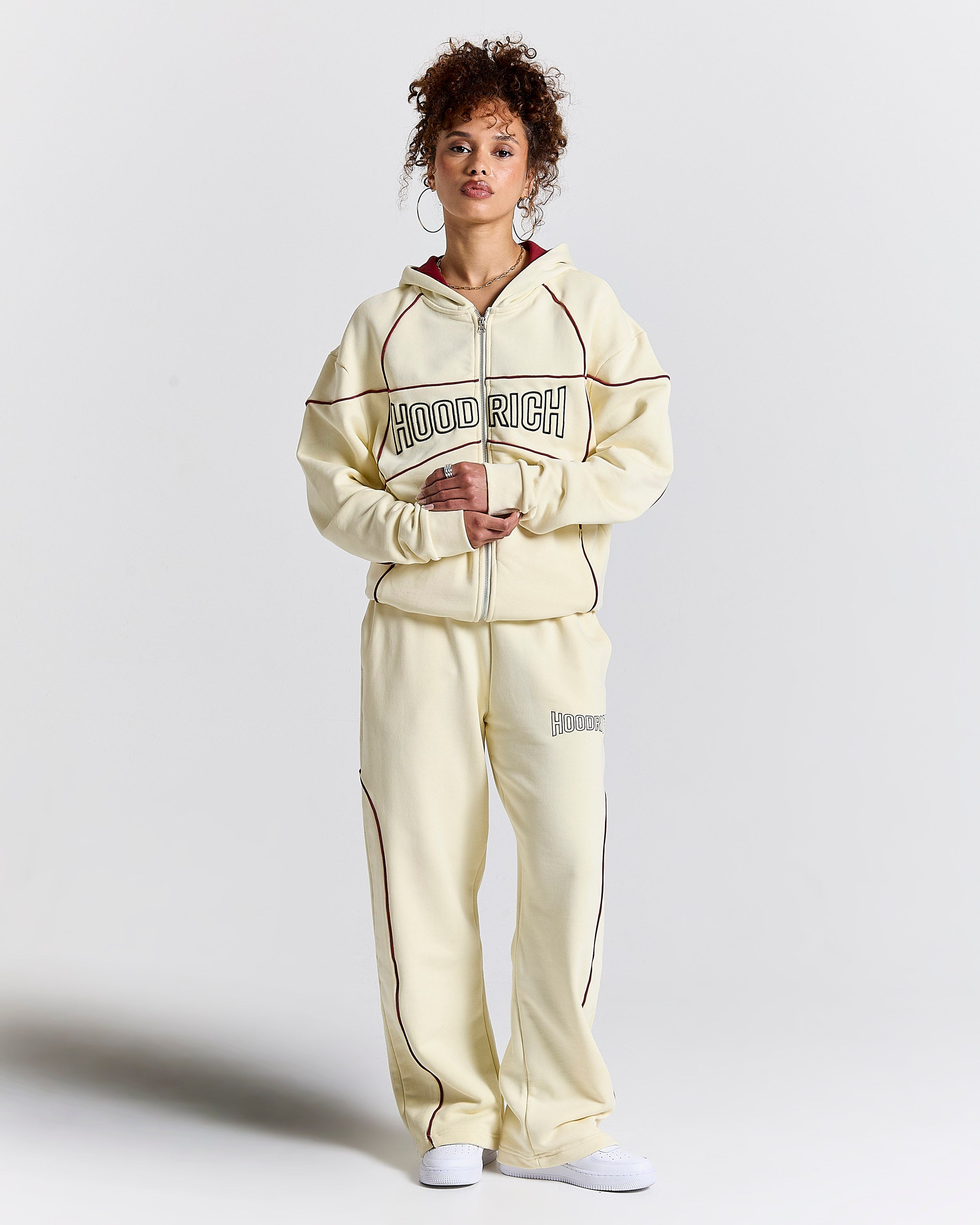 A model wearing a cream-colored hooded tracksuit with dark brown piping and 'HOODRICH' printed across the chest, standing against a light gray studio background.
