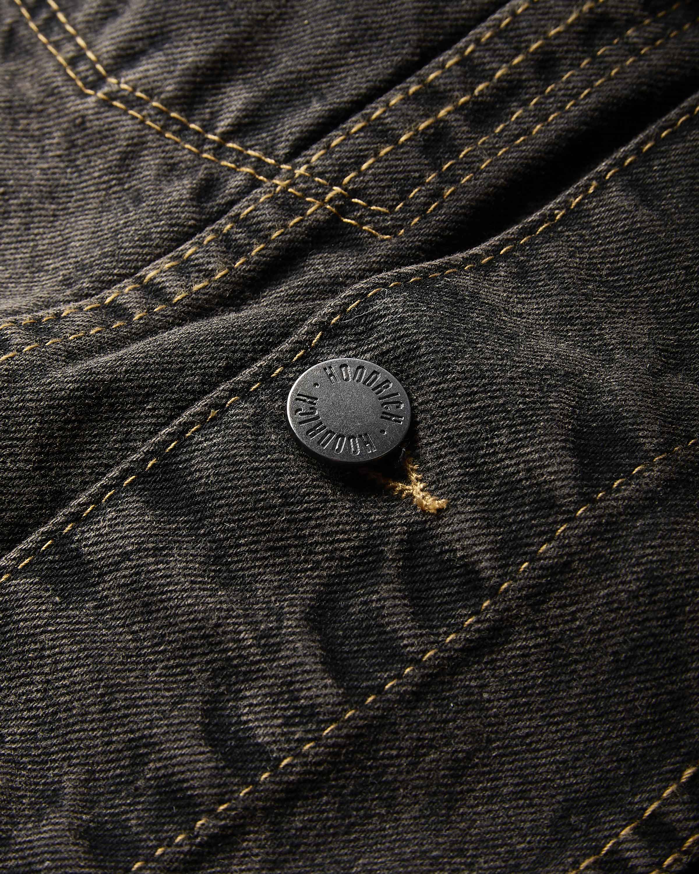 Close-up of dark charcoal gray denim fabric with contrasting tan stitching and a round black metal button embossed with 'HOODRICH'.