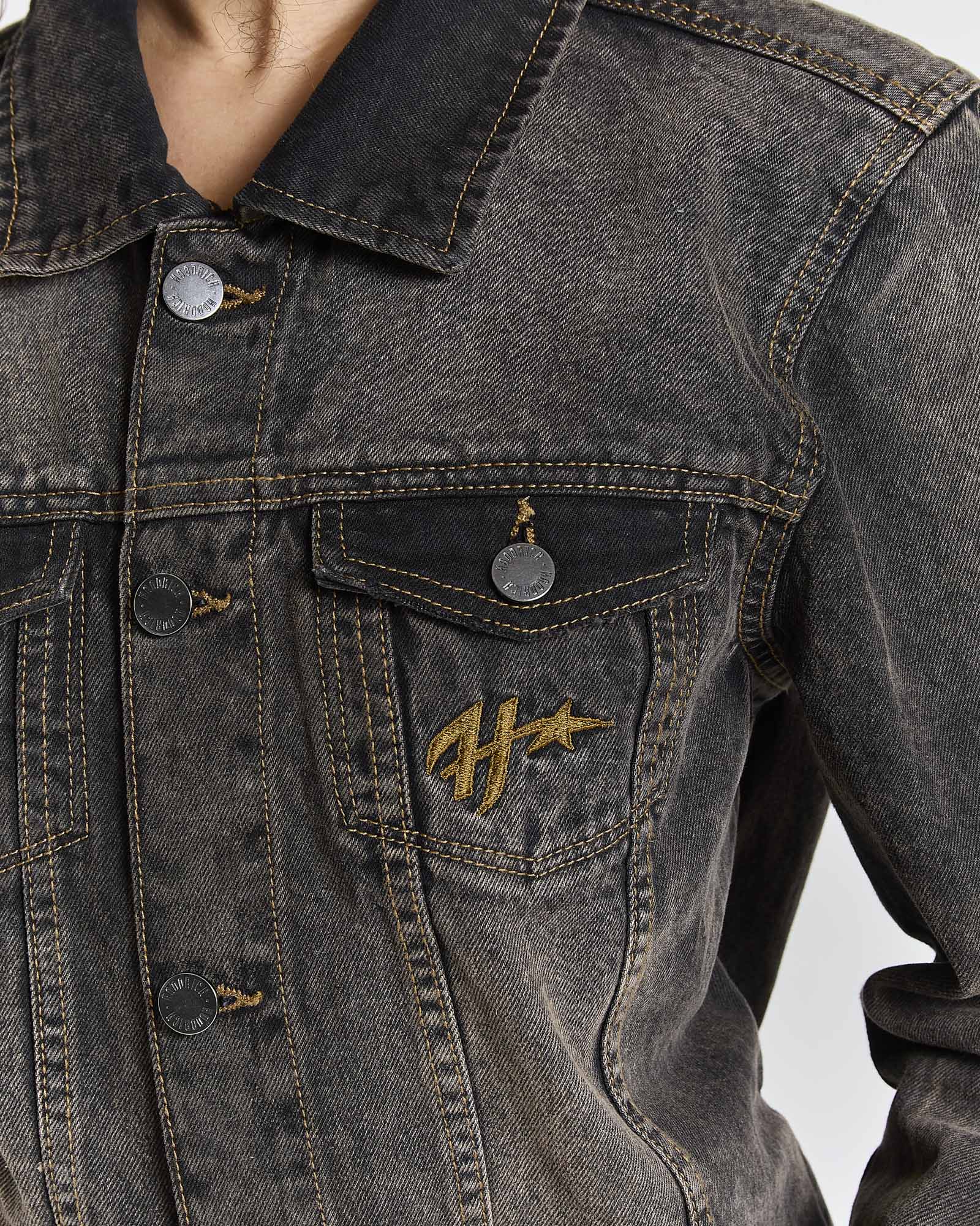 Close-up of a dark gray washed denim jacket with a gold embroidered logo on the chest pocket, featuring metal snap buttons and a collar.