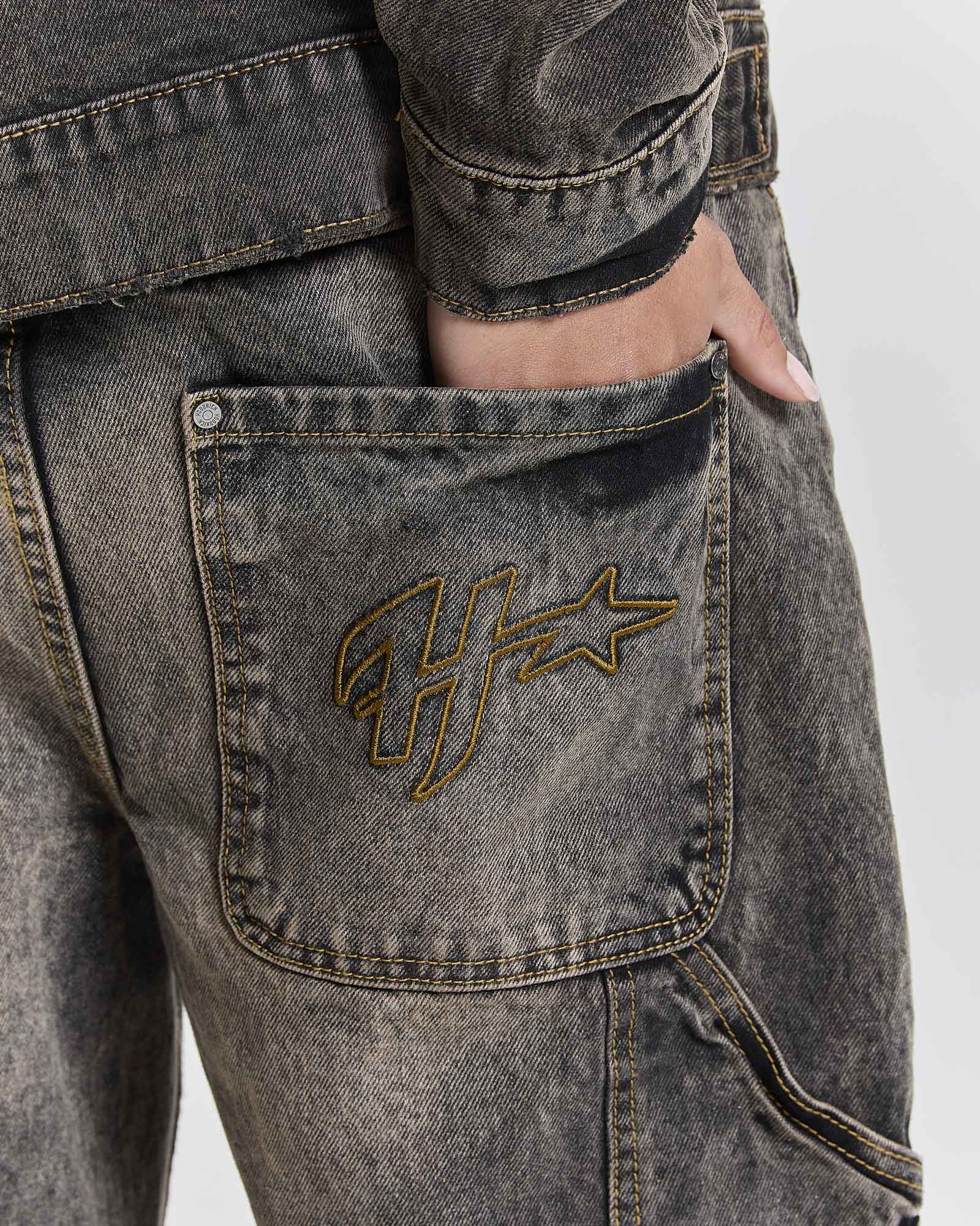 Close-up of a person wearing washed charcoal gray denim jeans with a gold embroidered Hoodrich  logo on the back pocket, showing a hand in the pocket.