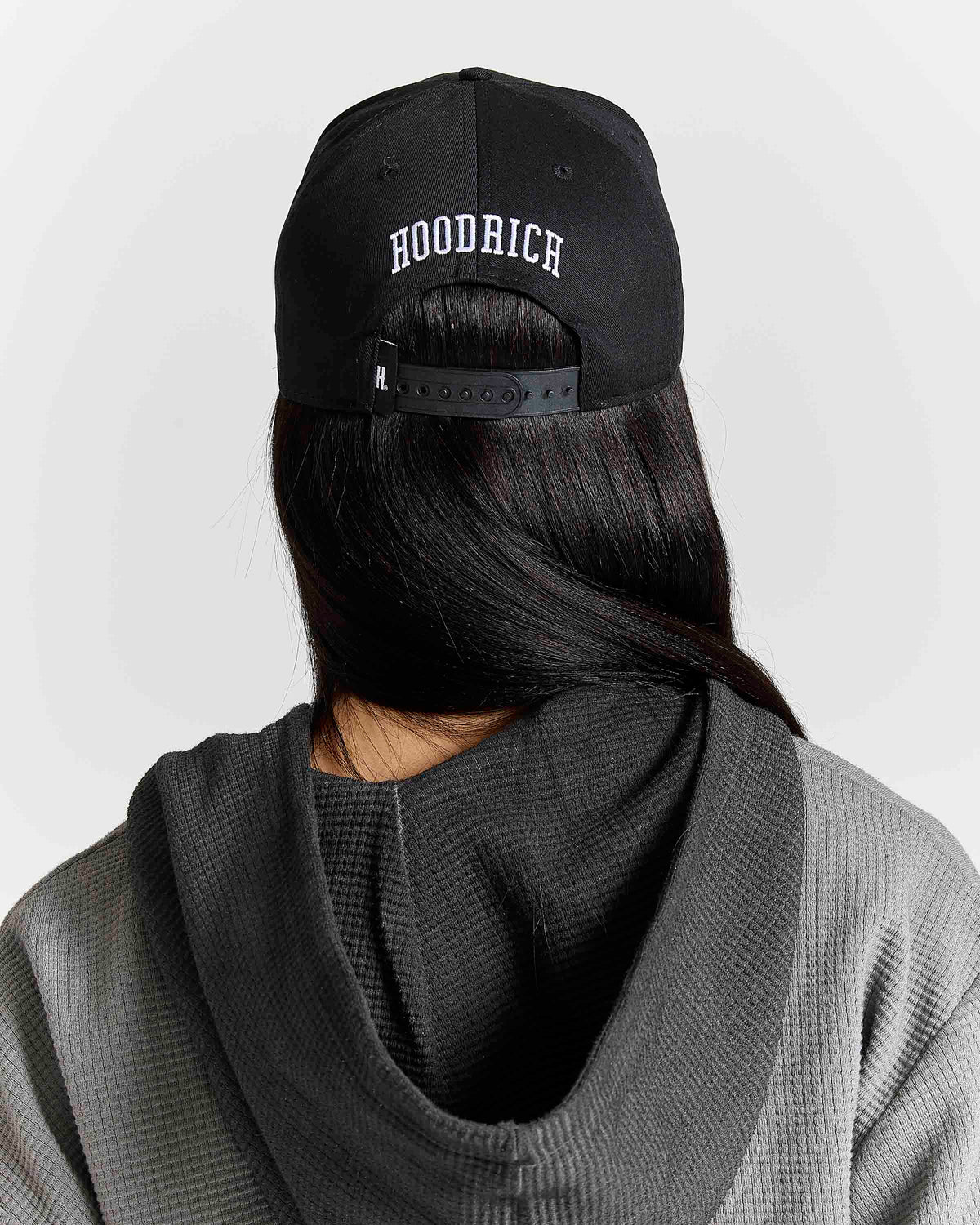A person shown from the back wearing a black baseball cap with 'HOODRICH' embroidered in white on the front, paired with a dark gray waffle-knit hoodie against a light gray background.