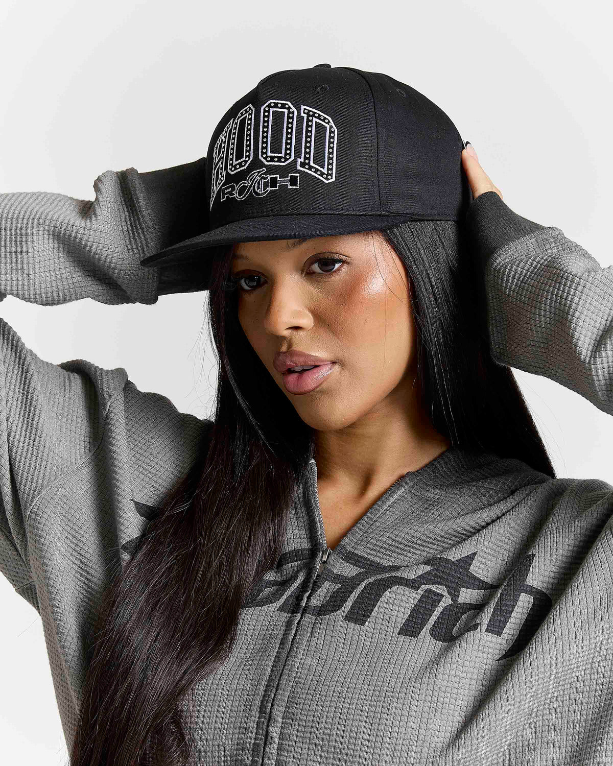 A woman wearing a black baseball cap with white 'HOODRICH' embroidery and a gray zip-up hoodie with 'earth' printed on the front, posed against a light gray background.
