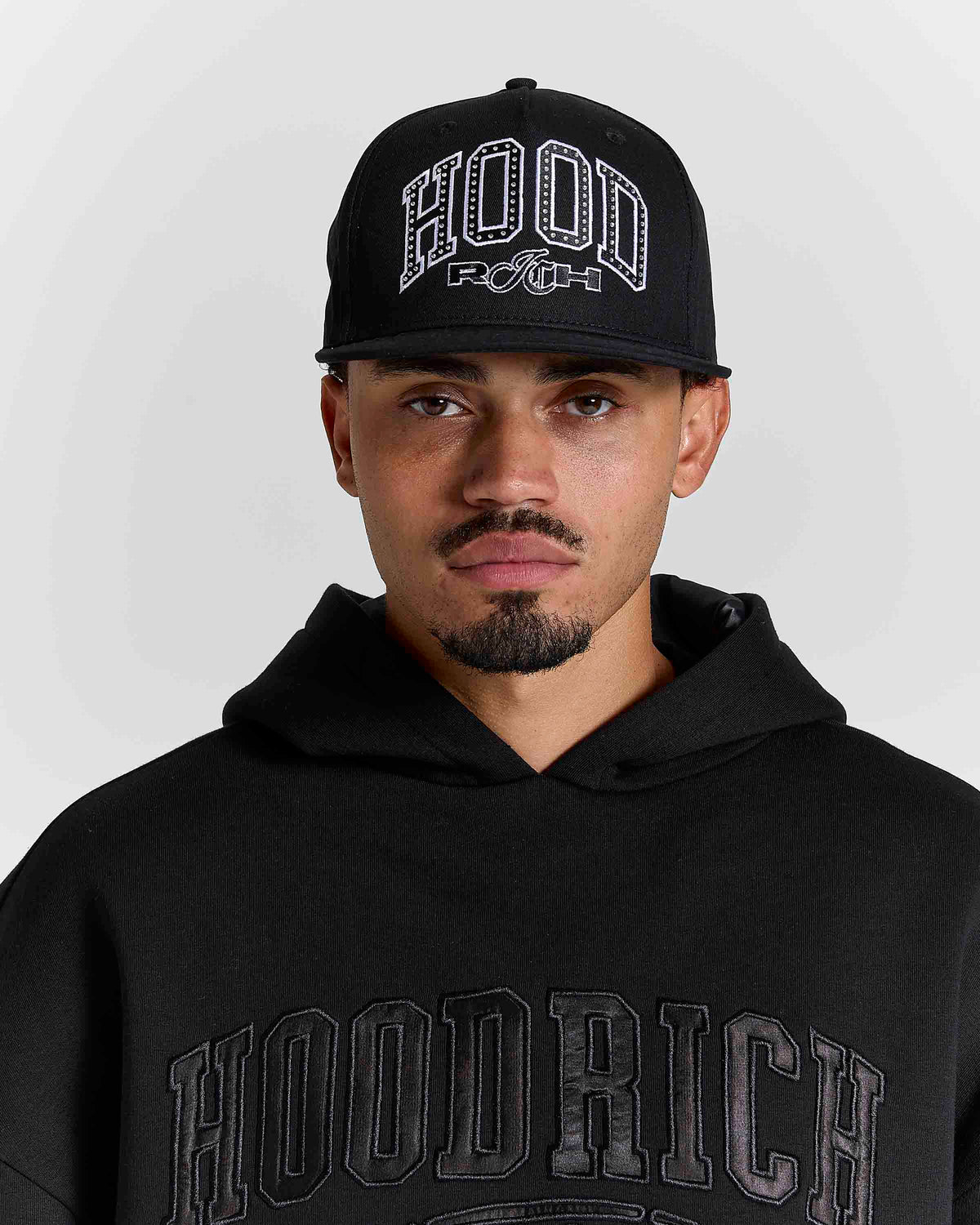 A man wearing a black HOODRICH baseball cap and a black HOODRICH hoodie, shown from the chest up against a light gray studio background.