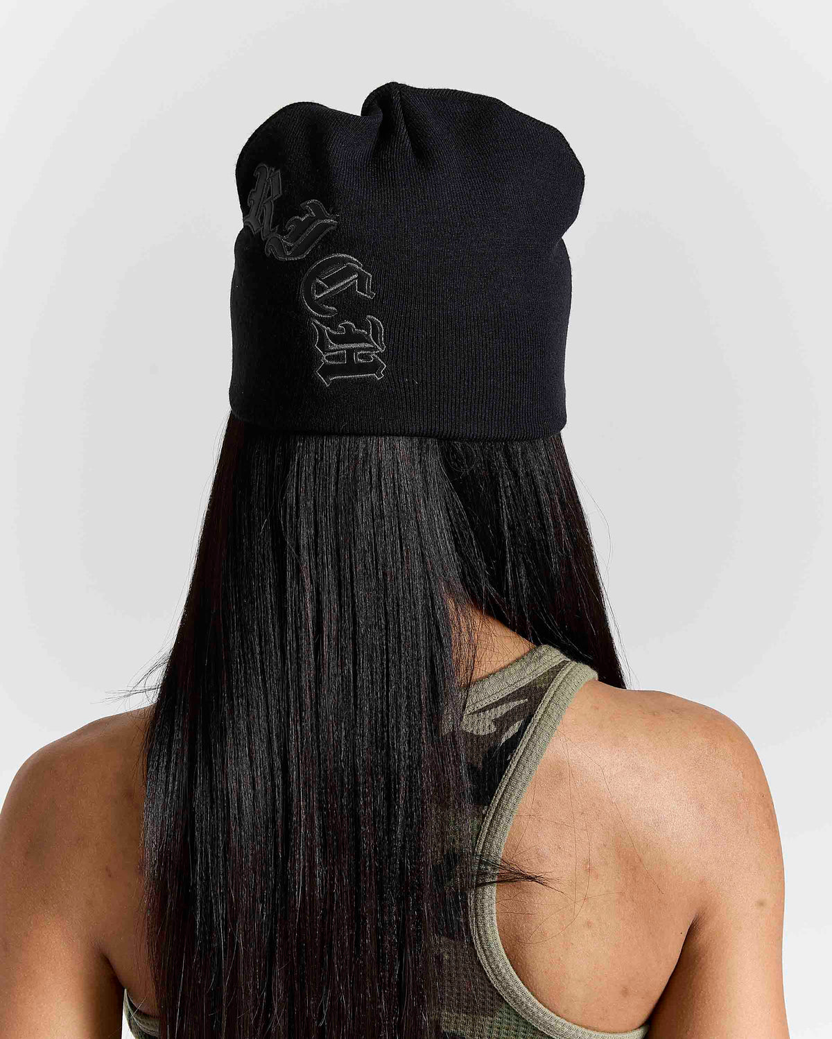 A person shown from the back wearing a black knit beanie with a tonal black embroidered logo on the side, against a light gray studio background.