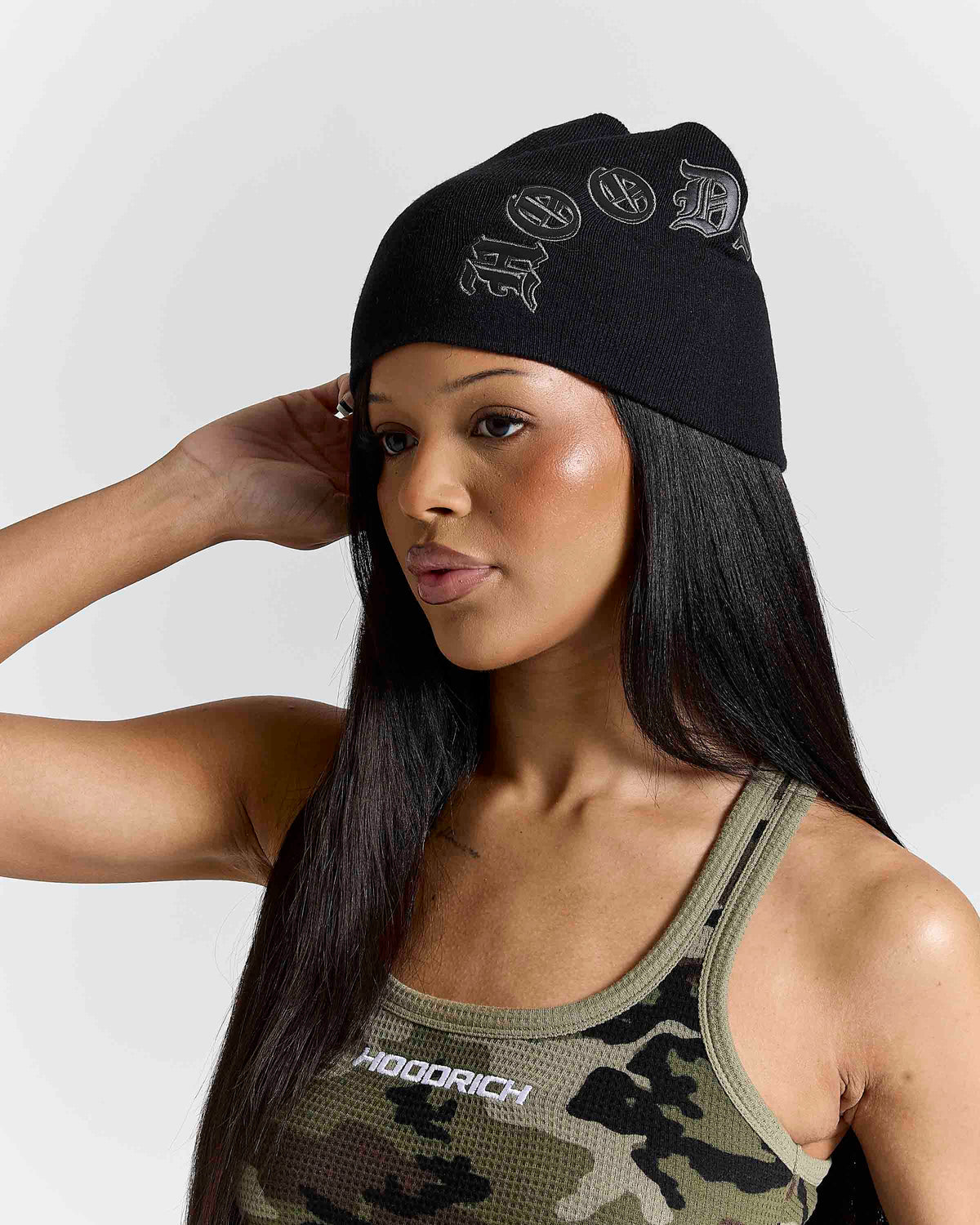 A woman wearing a black beanie with white 'HOODRICH' text and a camouflage tank top, posed against a light gray studio background.