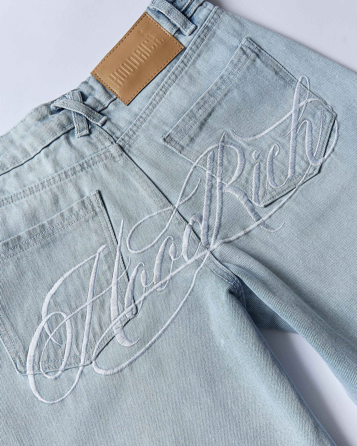 Light blue jeans with 'Hoodrich' embroidery on a white background