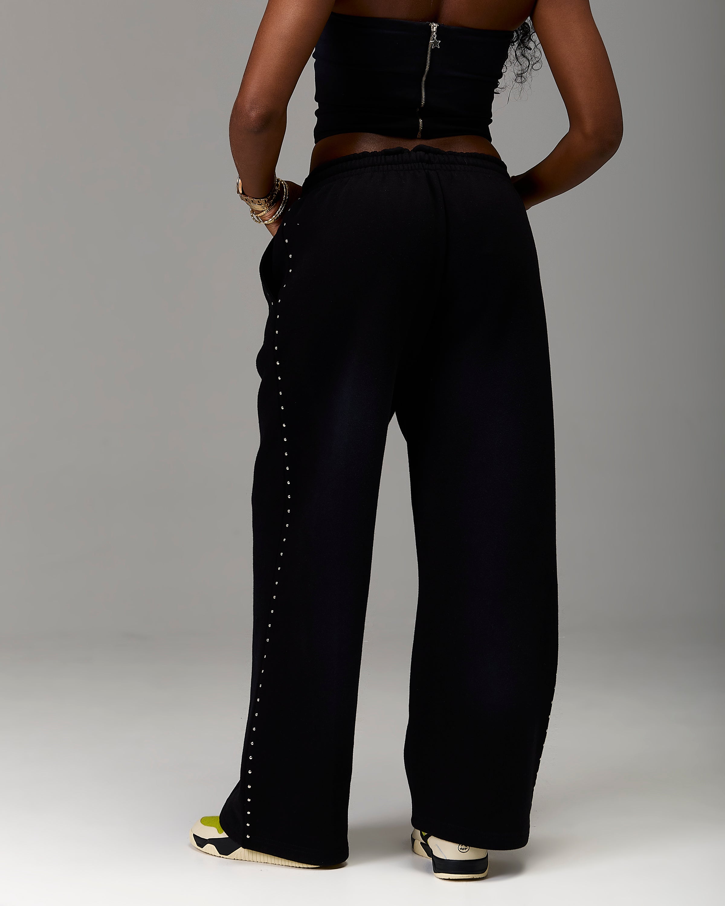 A model from the back wearing black wide-leg pants with a decorative line of silver studs down the side, paired with a black strapless top and sneakers, set against a plain gray studio background.